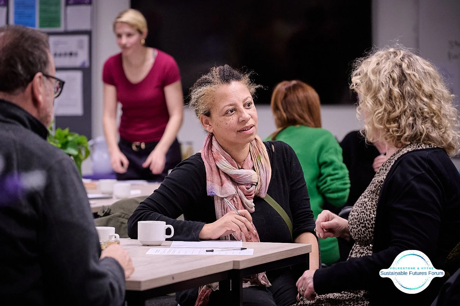 People engaged in conversation at a round table during the Sustainable Futures Forum, with cups and papers on the table and a woman standing in the background.