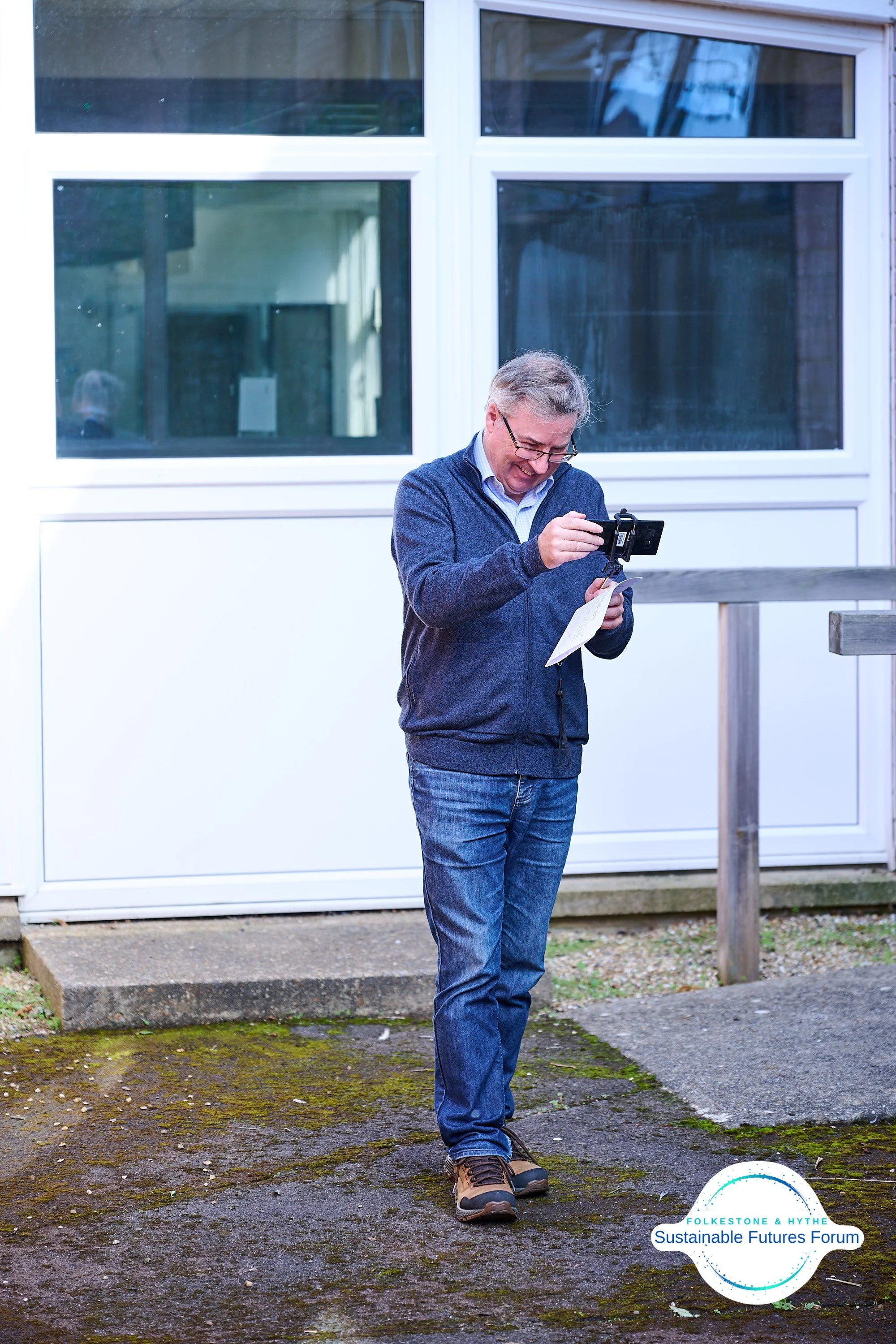 Man standing outdoors, smiling, holding a small camera or smartphone, with a notepad, wearing a blue jacket, jeans, and sturdy shoes, near a building with large windows and a moss-covered concrete surface, logo for Folketone & Hythe Sustainable Futur