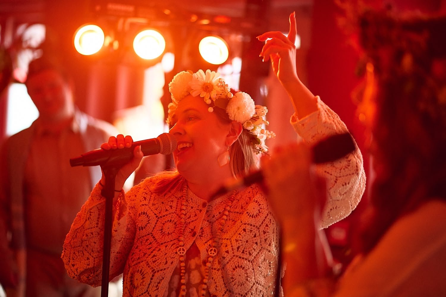 A woman singing into a microphone at a lively party or celebration, wearing a floral crown and lace clothing, with red and orange lighting.