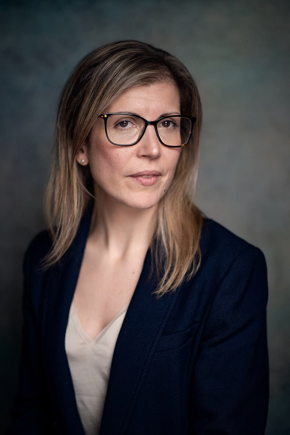 A woman with shoulder-length light brown hair, wearing glasses, a dark blazer, and a light-colored top, looking slightly to the side against a dark, blurred background.
