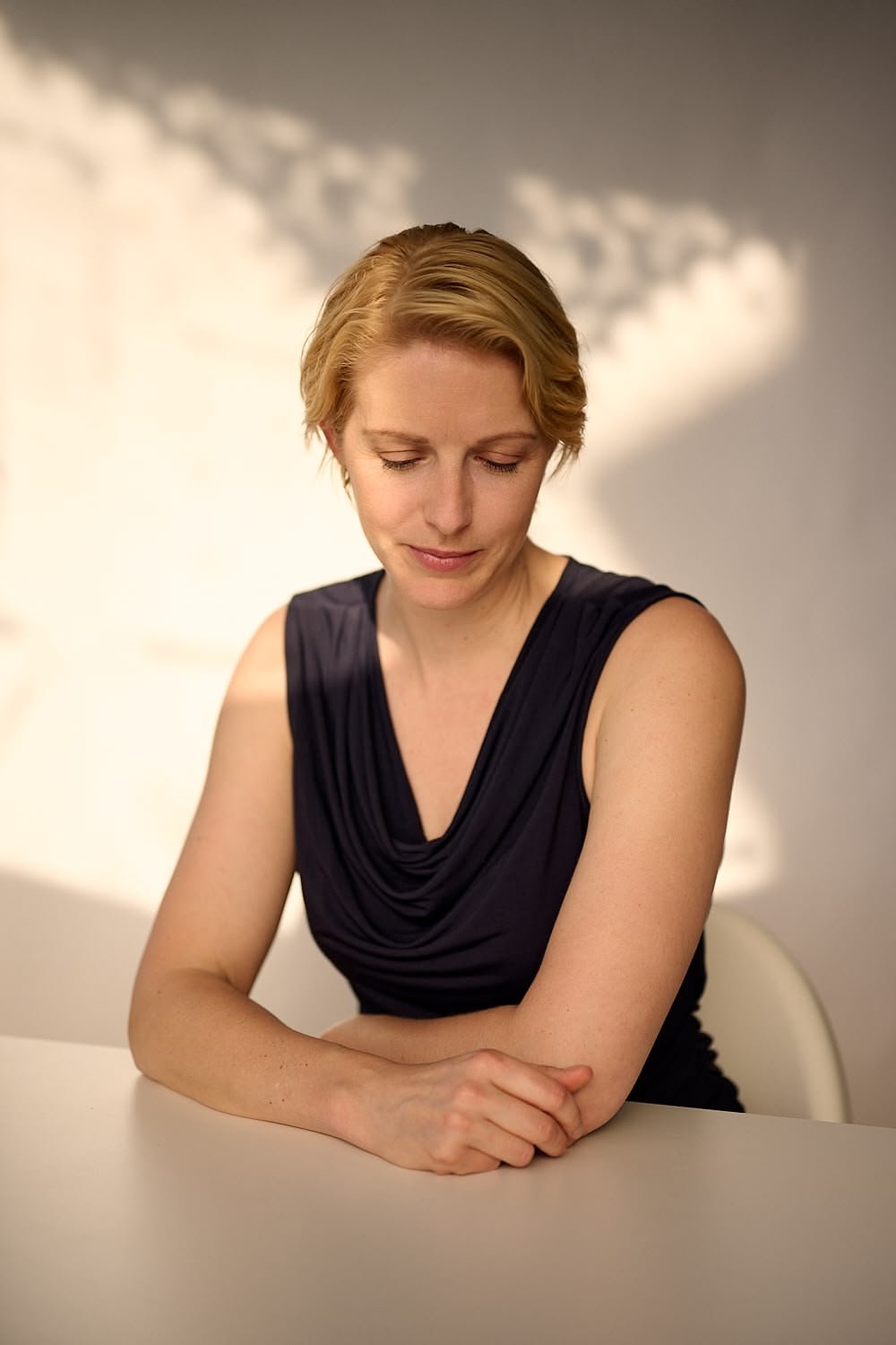 A woman with short blonde hair in a black sleeveless dress sitting at a white table, looking down with her arms crossed on the table, with a cloudy sky background.