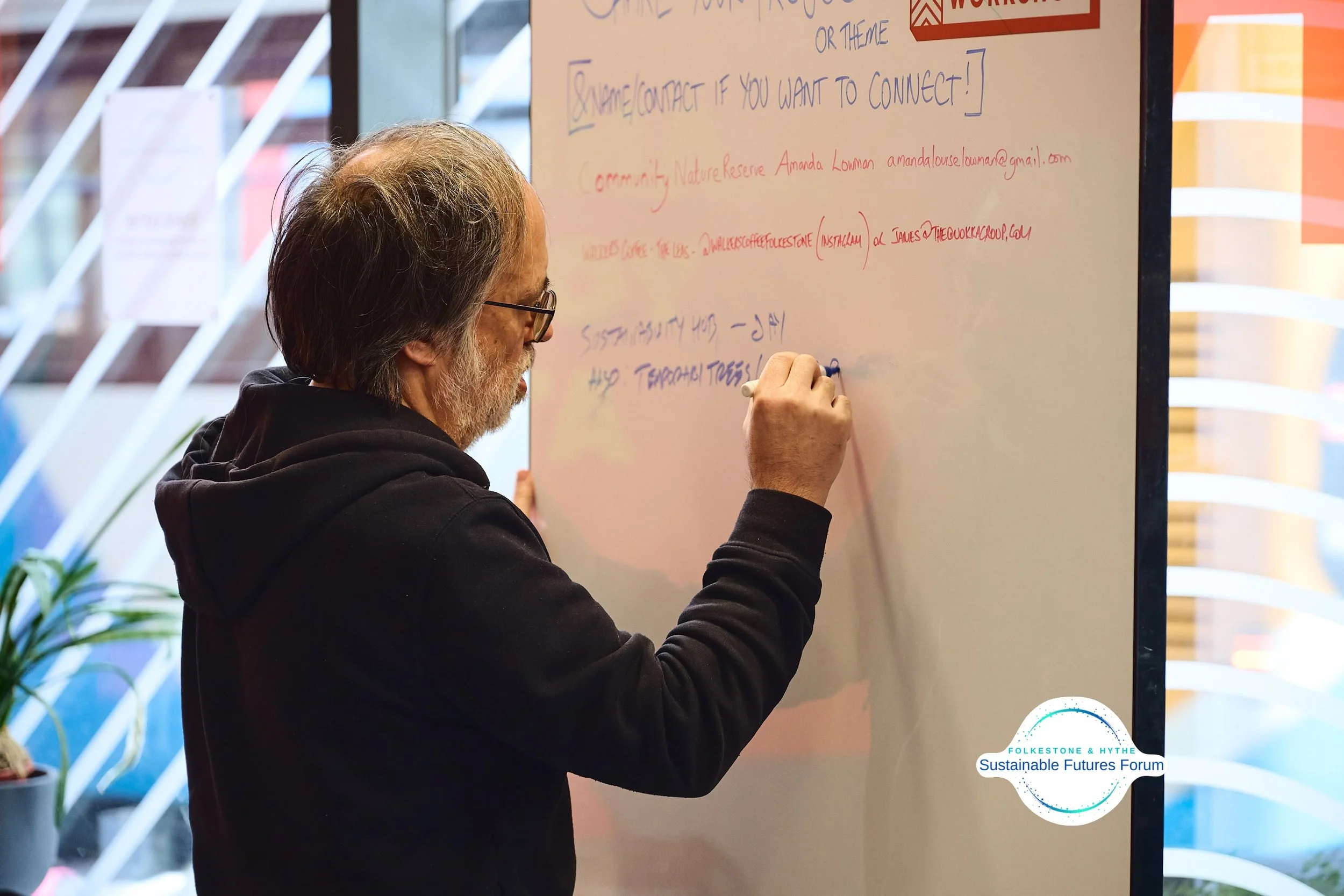 An older man with glasses writing on a large whiteboard in a meeting room, with notes and information about a community or nature reserve, and the logo of the Folkestone & Hythe Sustainable Futures Forum in the corner.