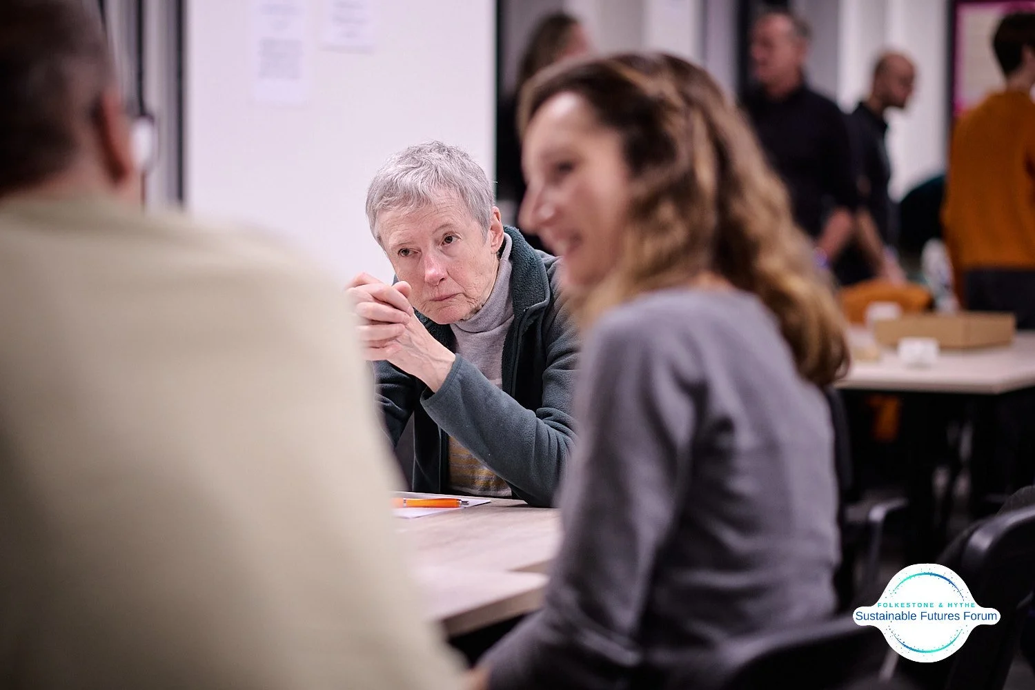 Older woman and two younger women sitting at a table engaged in a conversation during a forum or discussion event, with people in the background.
