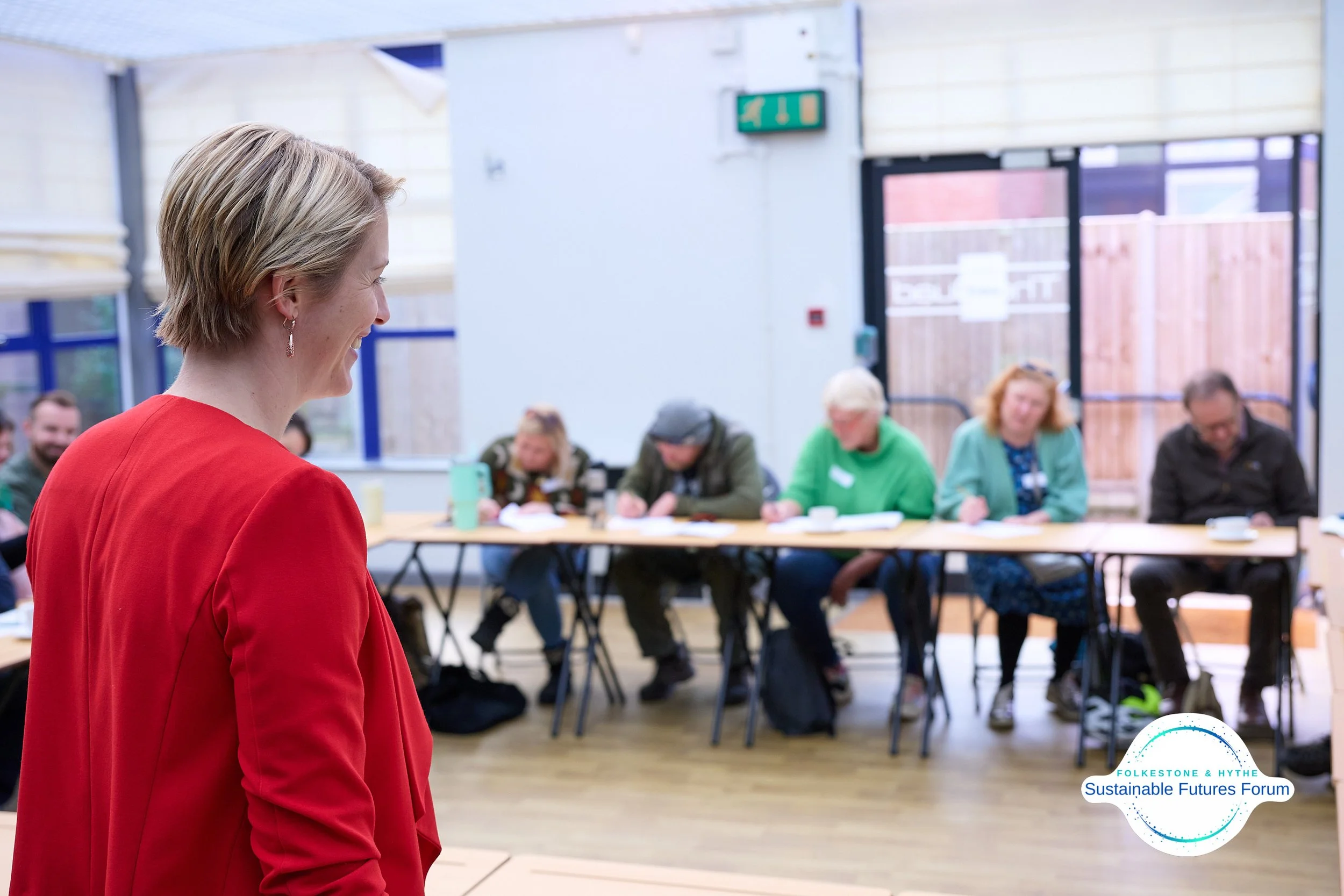 A woman in a red blazer smiling during a meeting with a group of people seated at a long table in a bright room with large windows and wooden floors. Logo reads "FolkesTone & Hythe Sustainable Futures Forum."