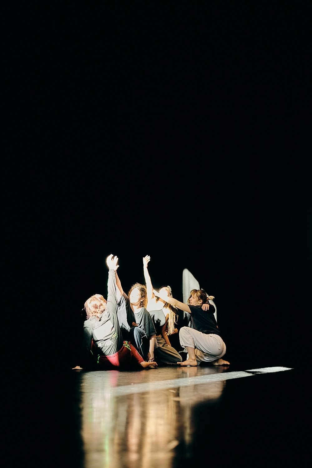 A group of people performing a dance or theater piece on a dark stage, illuminated by stage lighting.