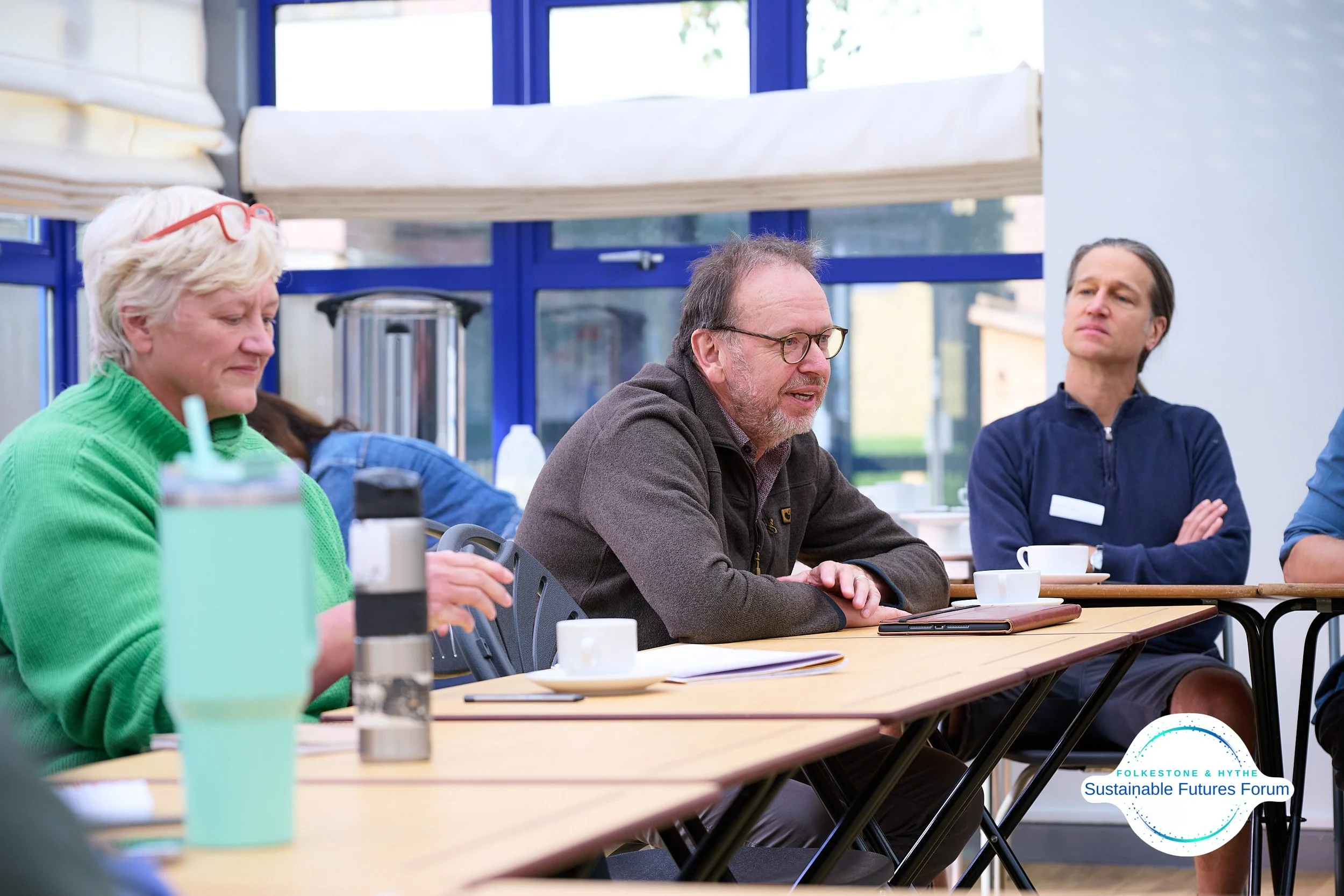 A group of people sitting around a table at a forum or conference, engaging in discussion. There are cups, notebooks, and a water bottle on the table. A logo for Folkesstone & Hythe Sustainable Futures Forum is visible in the bottom right corner.