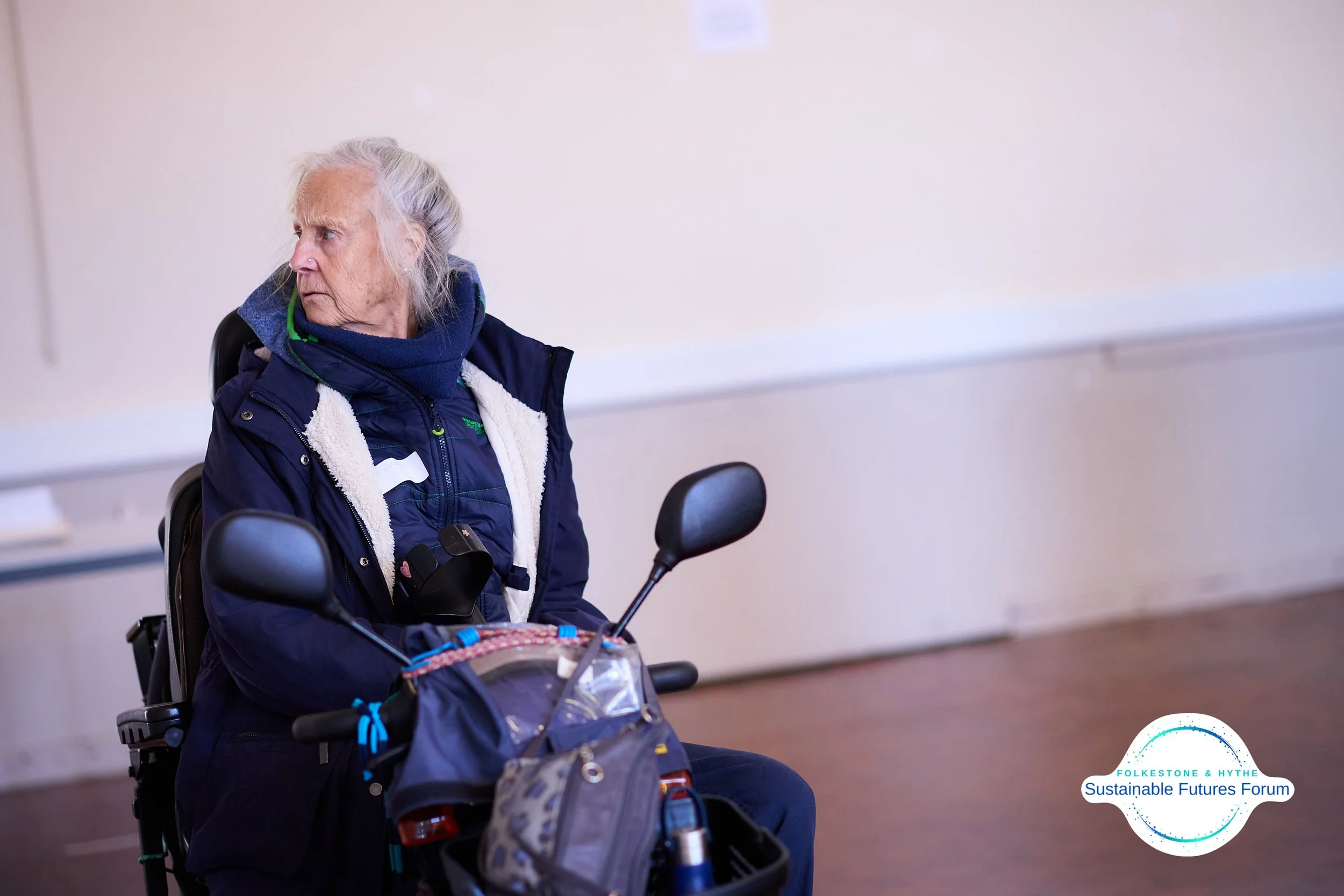 Elderly woman in a wheelchair looking to her left, wearing a blue jacket with white fleece lining and a navy blue hoodie, in a room with a white wall and a whiteboard in the background. There is a logo for the Folkestone & Hythe Sustainable Futures F