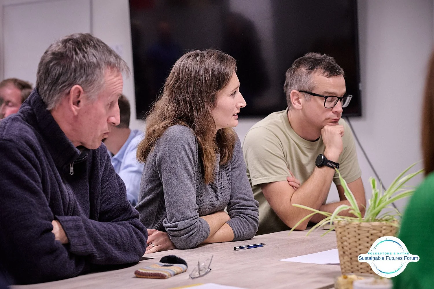 Three people sitting at a table during a discussion or meeting, with serious expressions. There is a potted plant and a logo at the bottom right corner that says "Sustainable Futures Forum".