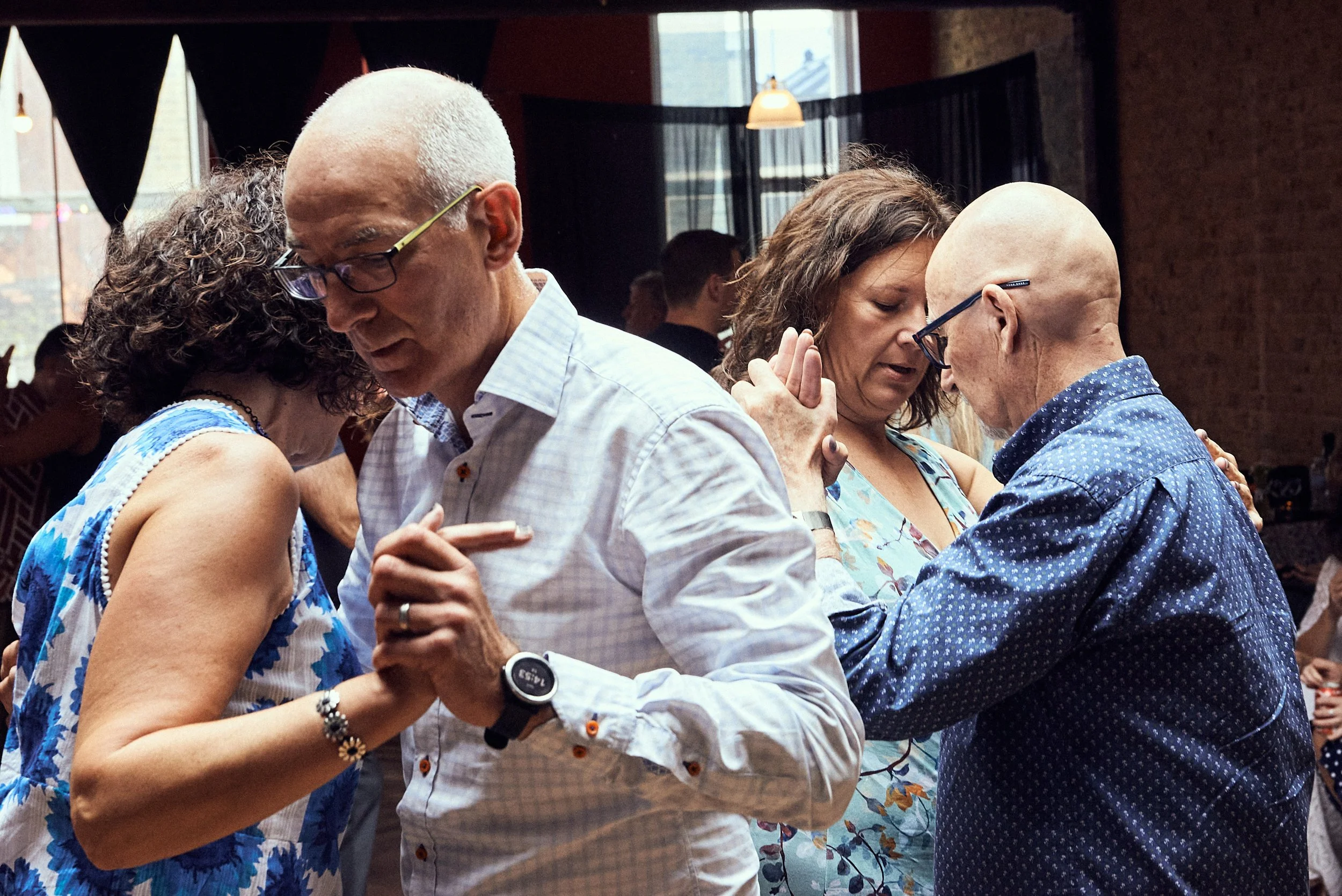Four people holding hands and dancing closely together in a social gathering indoors during daytime.