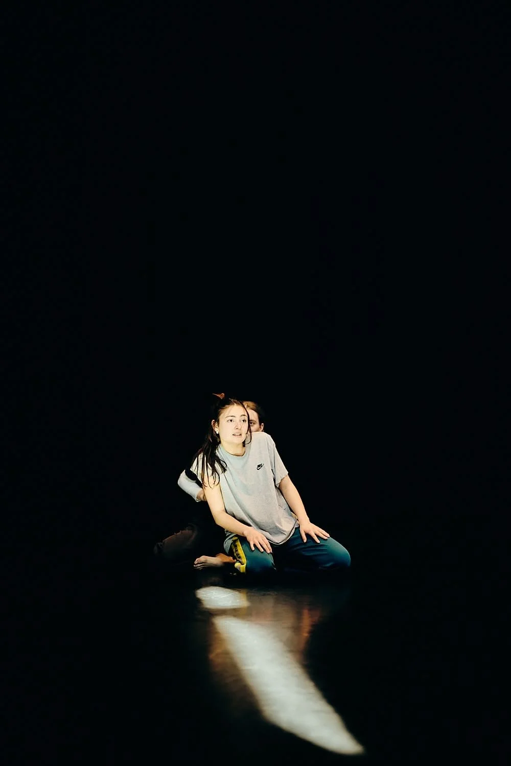 A young person with long dark hair, wearing a gray Nike t-shirt, kneeling on the ground in a dark space with a spotlight reflecting on the floor.