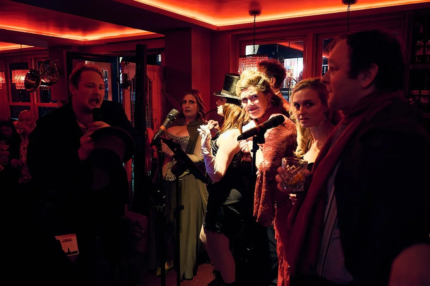 A group of people gathered in a dimly lit room, some holding drinks. A man on the left is speaking into a microphone, wearing a black suit and holding a hat. Several women with elaborate costumes, including a woman with a black hat, are standing behi