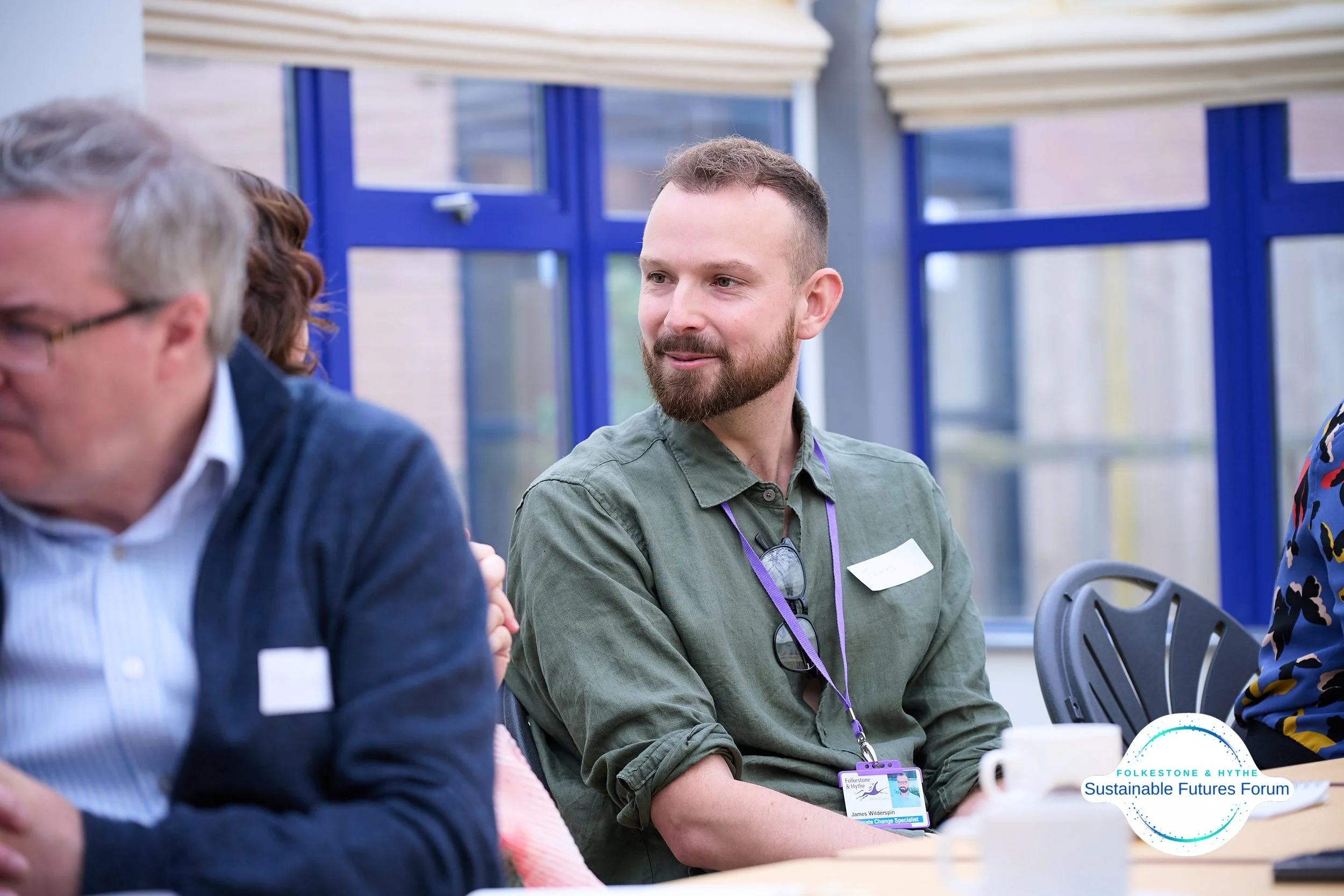 A man with a beard and short hair, wearing a green shirt and glasses, sitting at a table during the Folkesone & Hythe Sustainable Futures Forum, smiling and looking to the right.