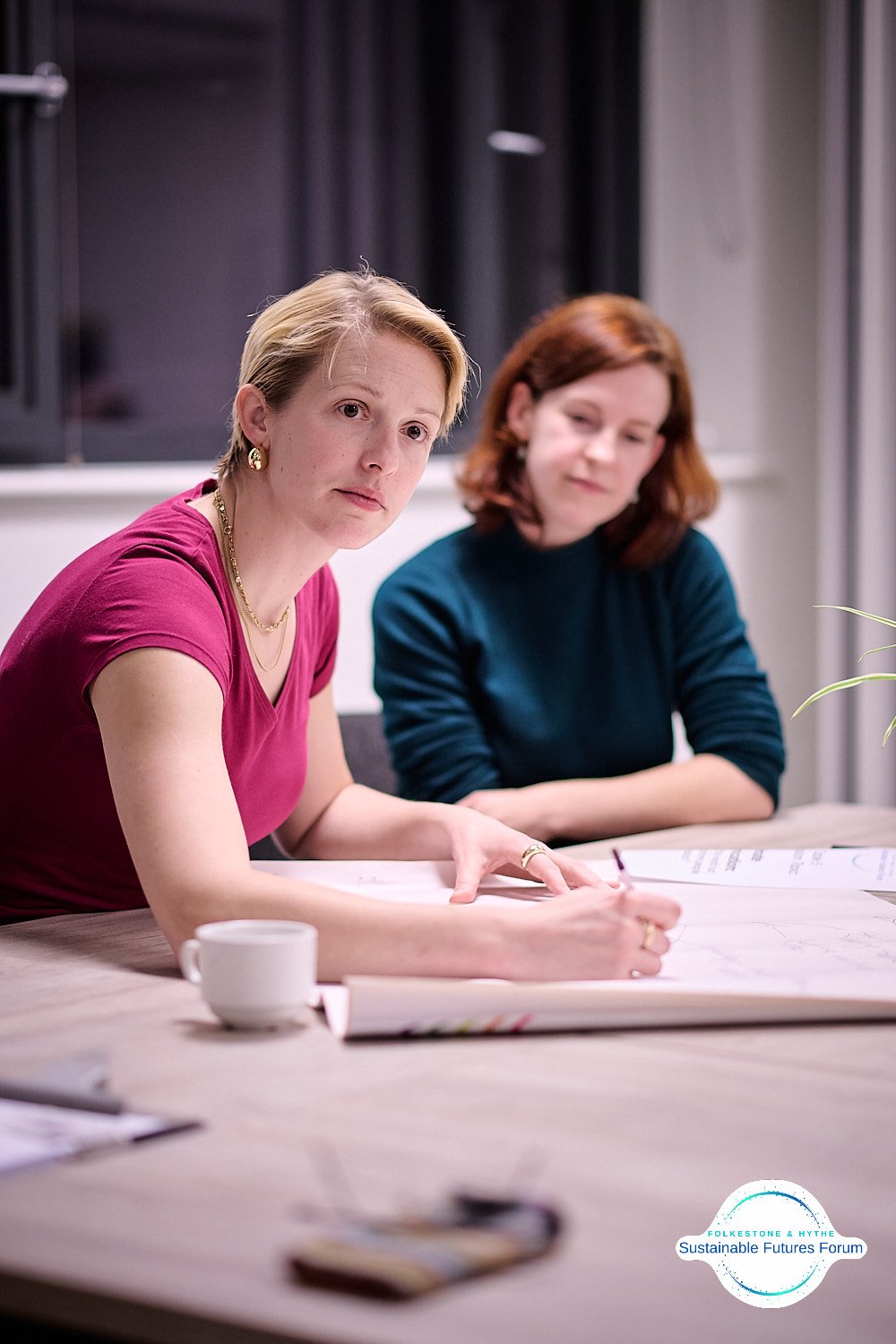Two women sitting at a table during a meeting, one appears to be explaining something on a large sheet of paper, with a coffee cup and other papers on the table, in a room with black curtains and a window in the background.