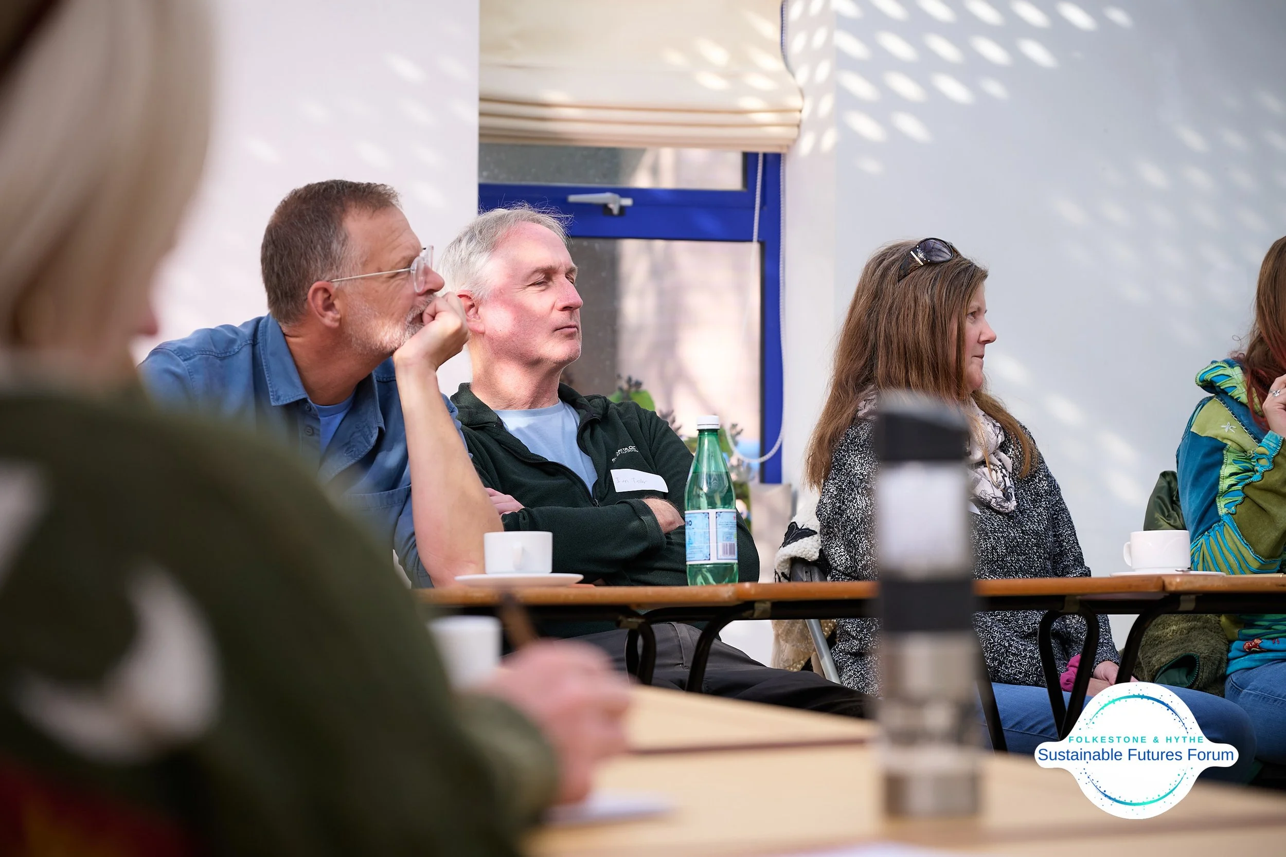 People attentively listening at a forum, seated at a table with cups and water bottles, during the FolkeStone & Hythe Sustainable Futures Forum.