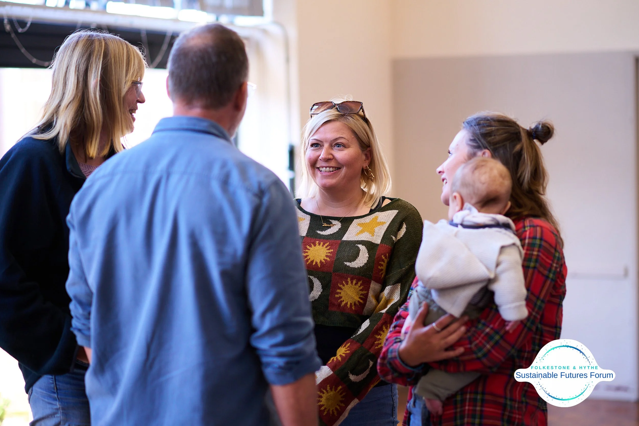 Group of people talking and smiling at an indoor event, with a logo for FolkesTone & Hythe Sustainable Futures Forum in the bottom right corner.