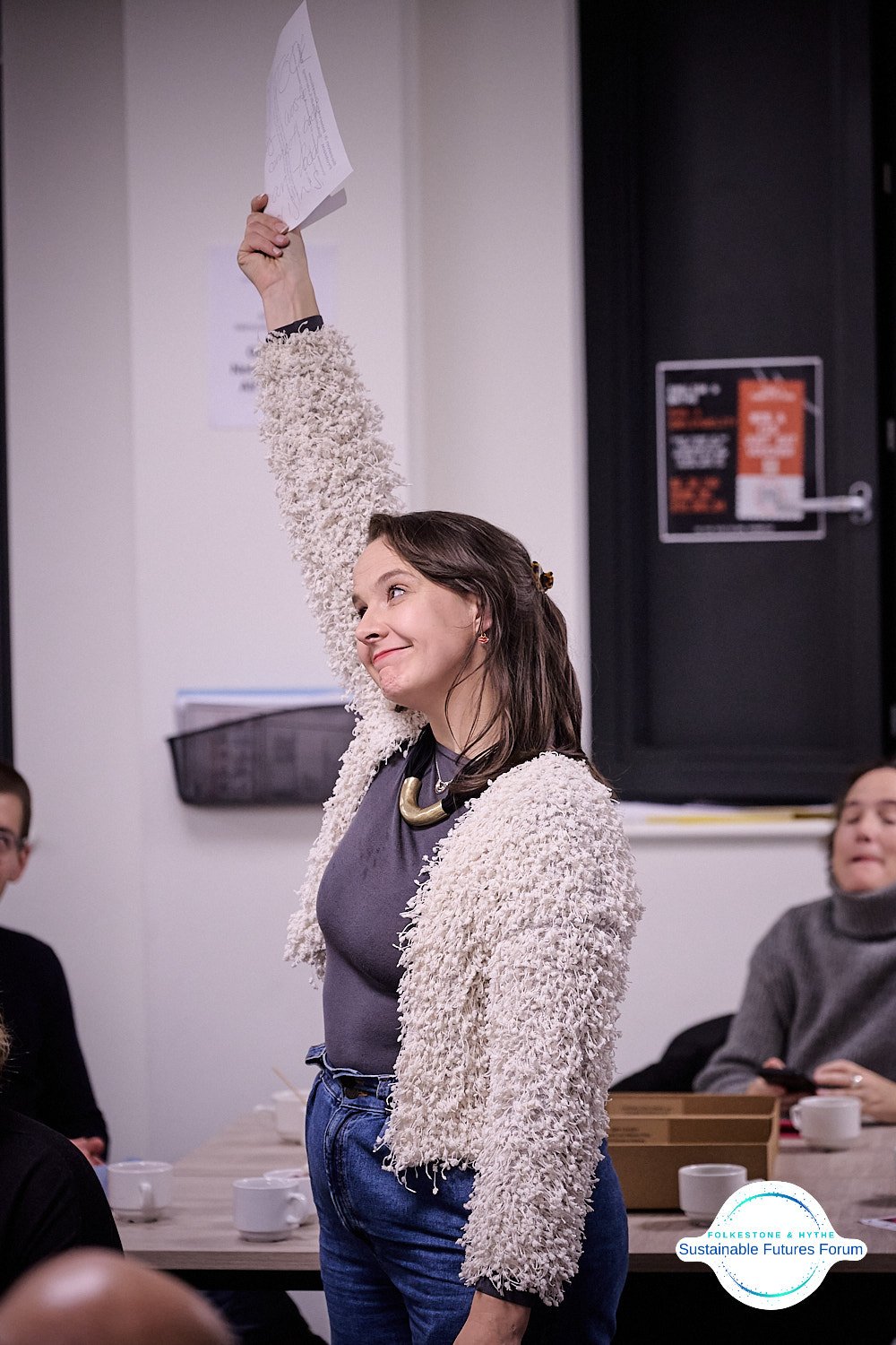 A woman with shoulder-length brown hair, wearing a gray shirt and a cream-colored textured jacket, raises her hand holding a piece of paper at a meeting or discussion, with other people seated around her. She has a cheerful expression.