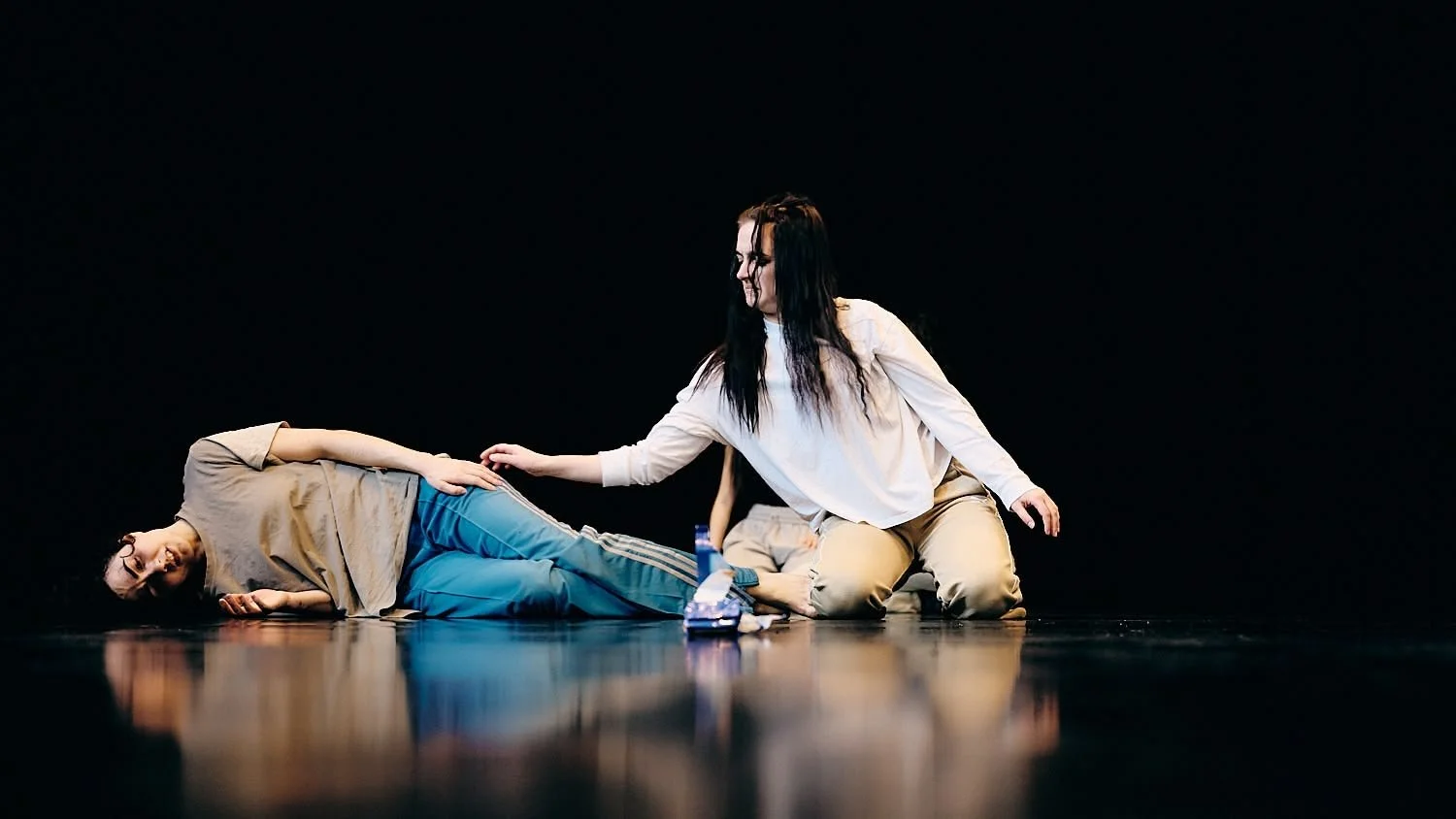 Two women on stage, one lying on the floor and another kneeling, engaging in an acting scene. The woman on the floor is wearing a beige shirt and blue pants, while the woman kneeling is wearing a white shirt and beige pants, reaching out to the other