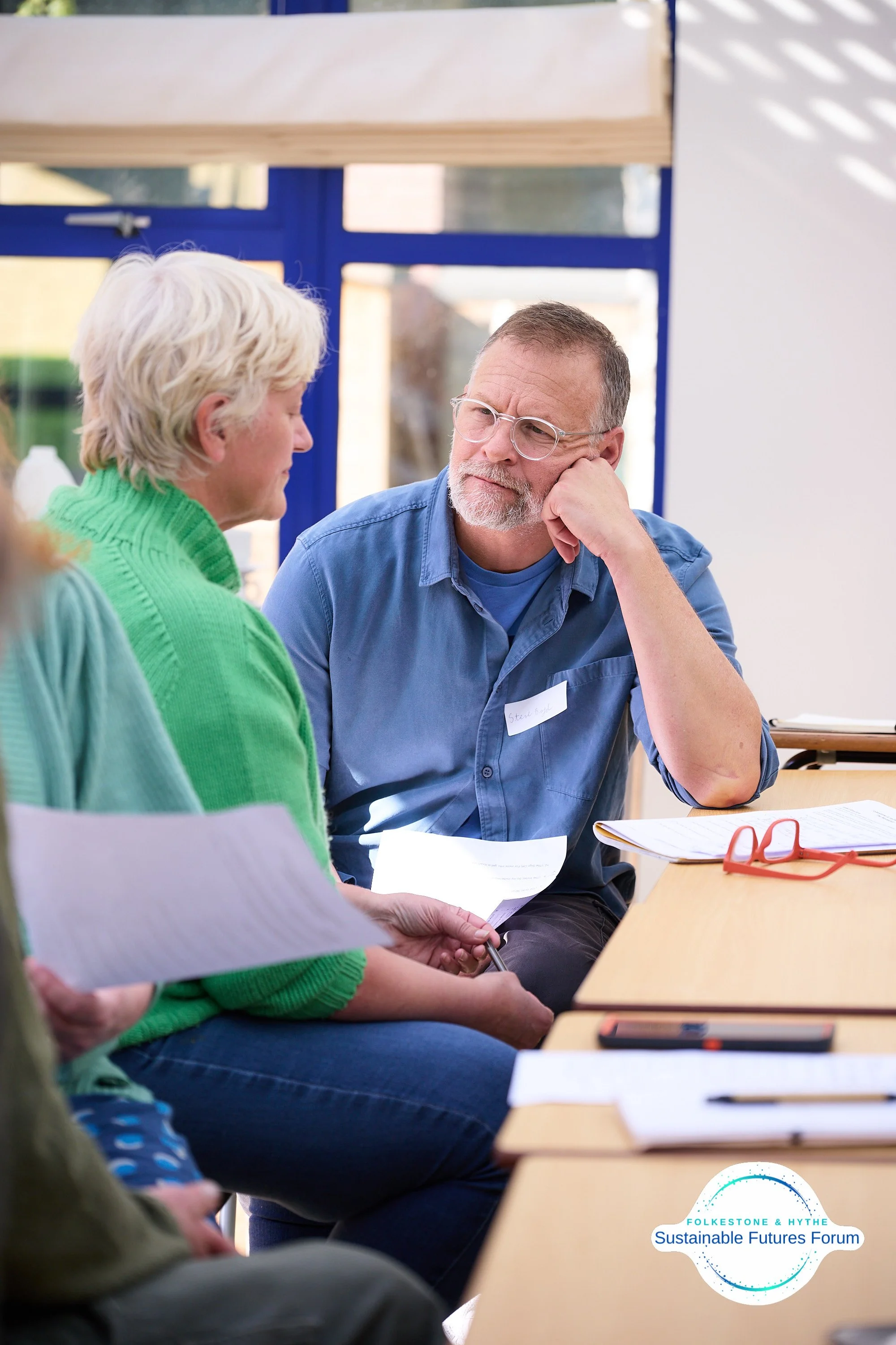 A middle-aged man with glasses and a beard, wearing a blue shirt, is engaged in conversation with an older woman in a green sweater, at a table during a community or forum event. There are papers, a pair of red glasses, and a badge on the man, with t