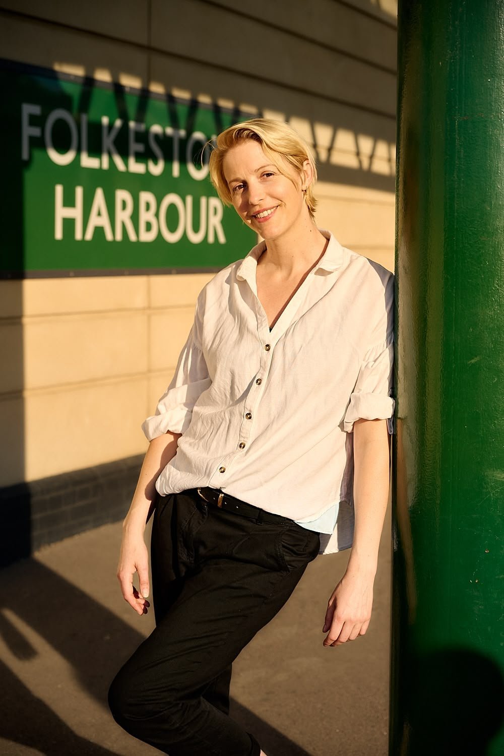 Woman with short blonde hair, white button-up shirt with rolled-up sleeves, and black pants, leaning against a green pole near Folkestone Harbour sign, during golden hour sunlight.