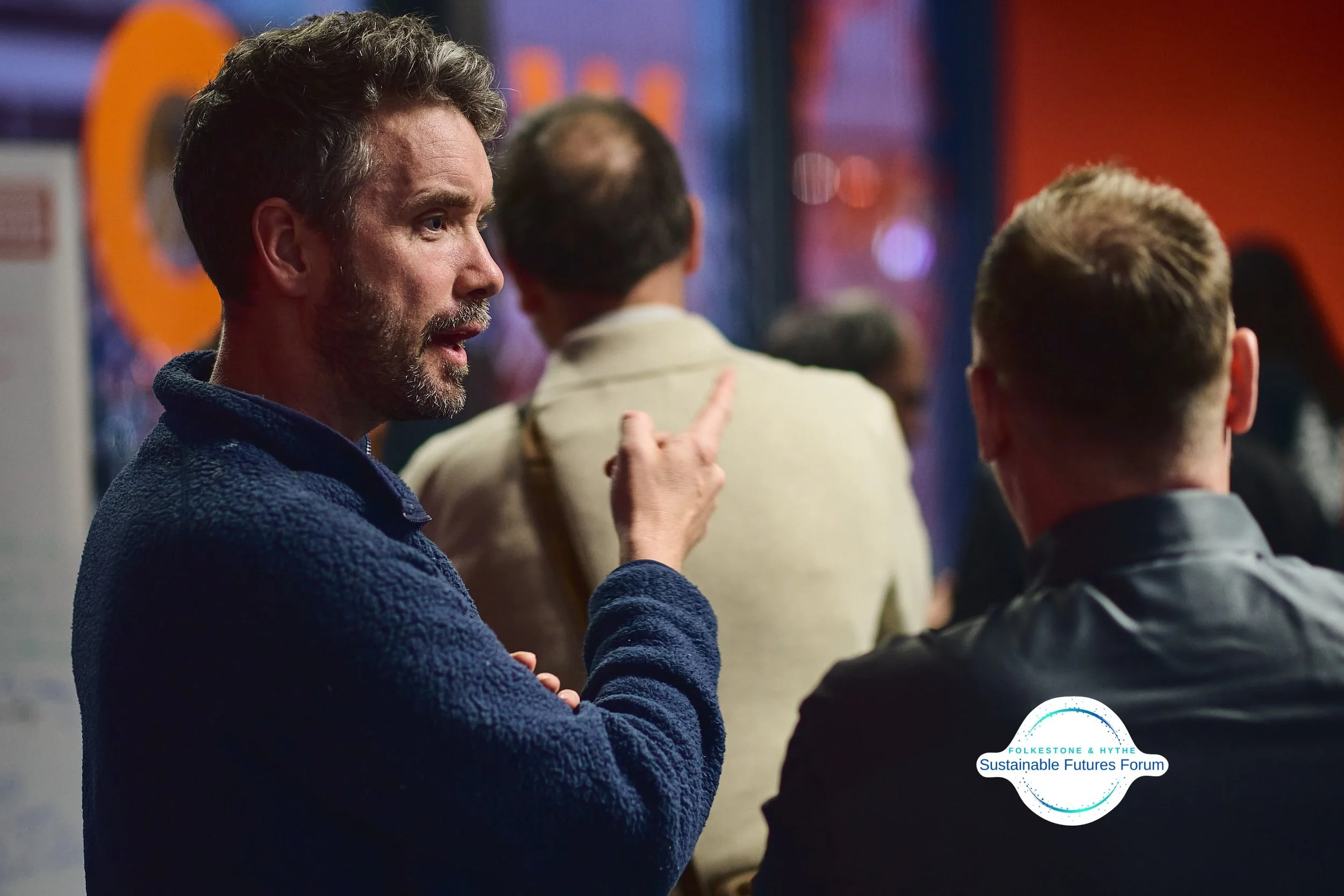 A man with gray hair and a beard, wearing a blue textured jacket, is pointing and speaking to another man with blonde hair and a black leather jacket at what appears to be a social event or forum. There are other people in the background. A logo in t