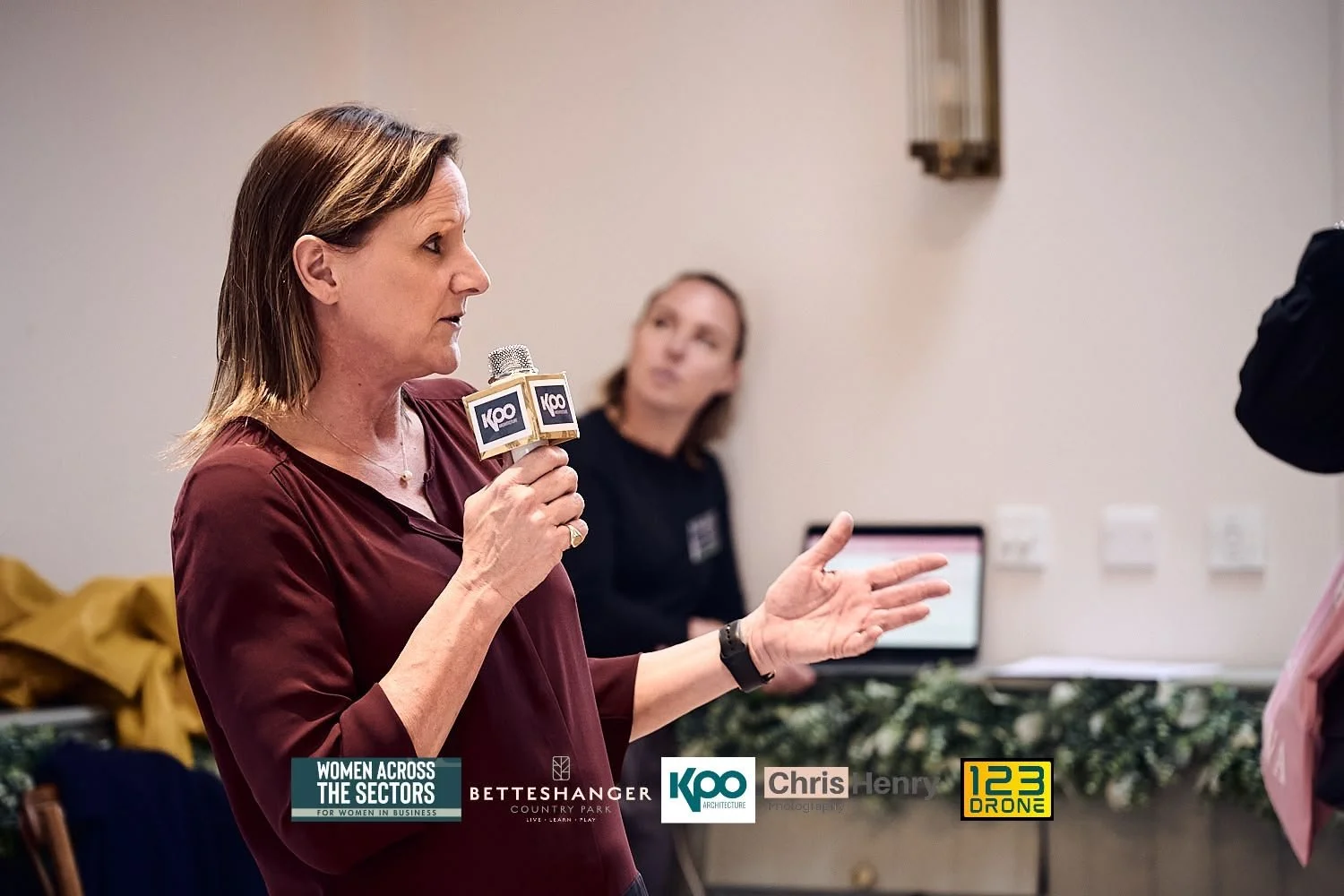 A woman speaking into a microphone with a 'KCO' logo, gesturing with her right hand during a discussion at a professional event, with another woman listening in the background.