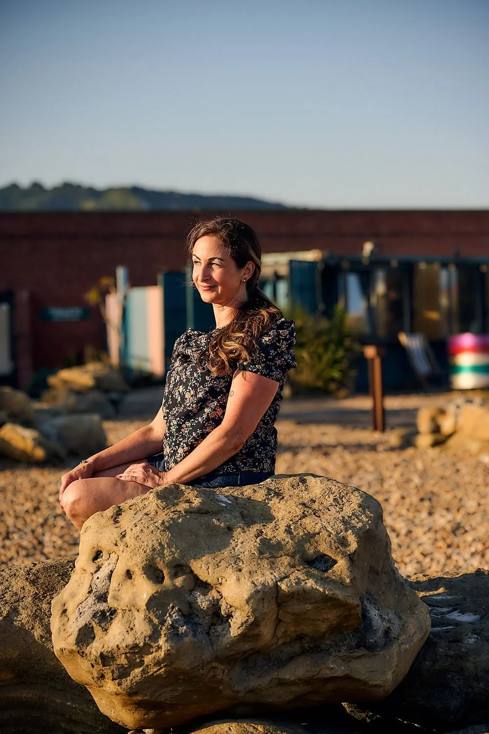 A woman with long curly hair, wearing a dark floral shirt, sits cross-legged on a large rock at the beach during golden hour, with a background of pebbles, rocks, and a building with a brick wall.