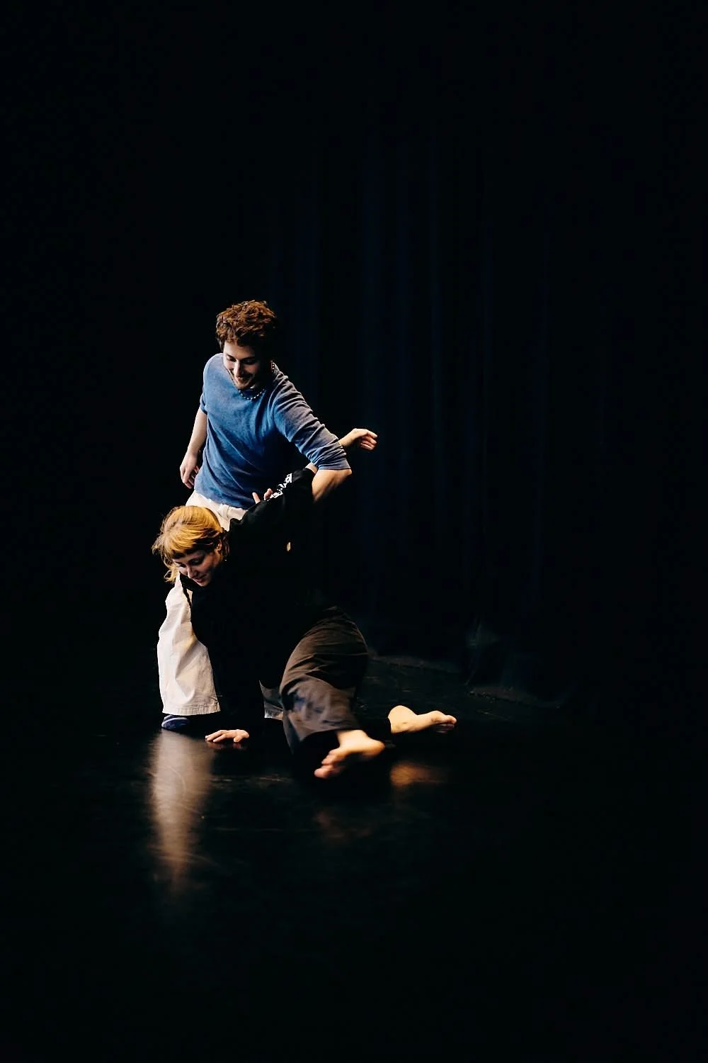 Two performers engaged in a dance or theatrical performance on a dark stage, one standing and the other on the ground, both dressed casually.