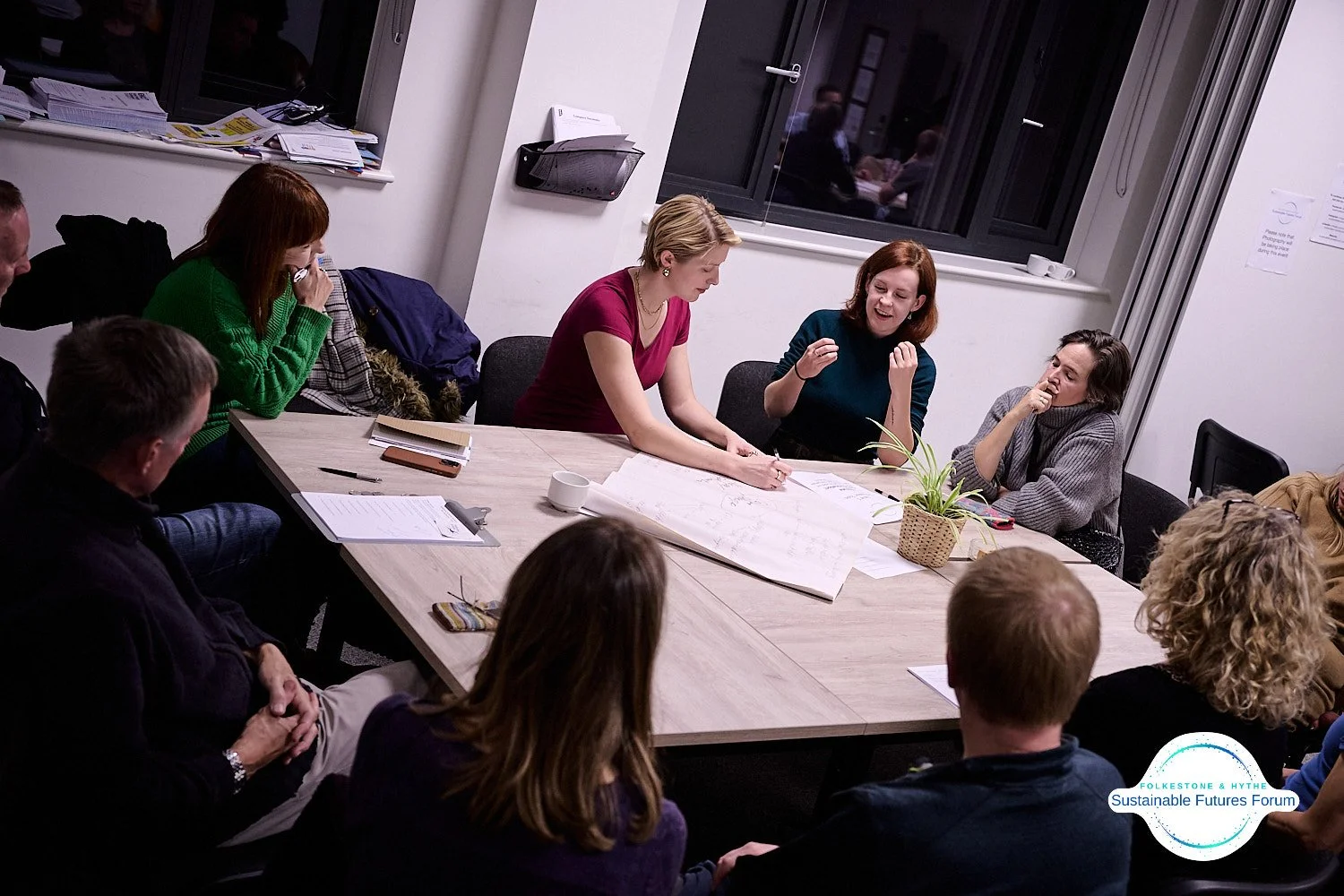 Group of people in a meeting room having a discussion around a table, with some taking notes and others talking, with documents and a plant on the table.