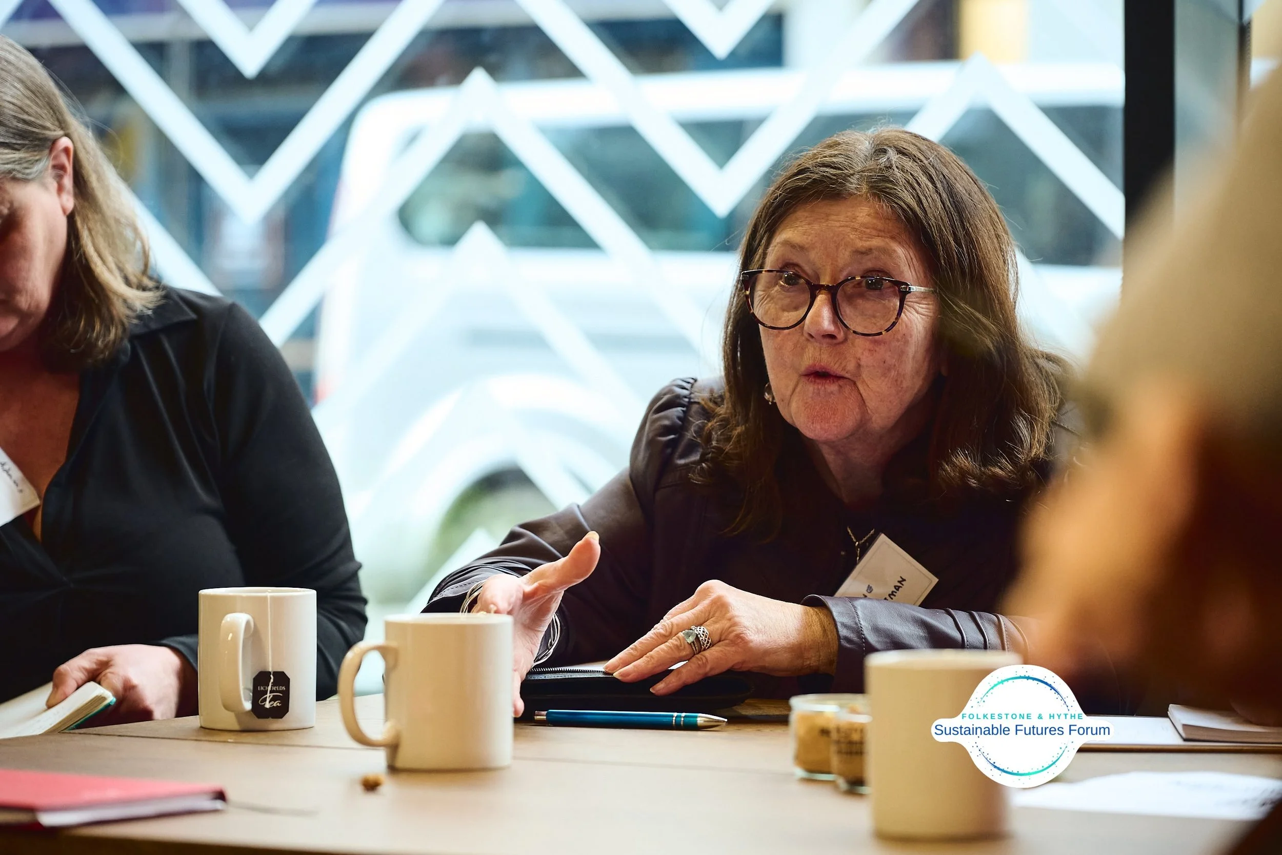 Woman with glasses speaking during a meeting at a table with cups, notebooks, and pens, with a window featuring a geometric pattern in the background.