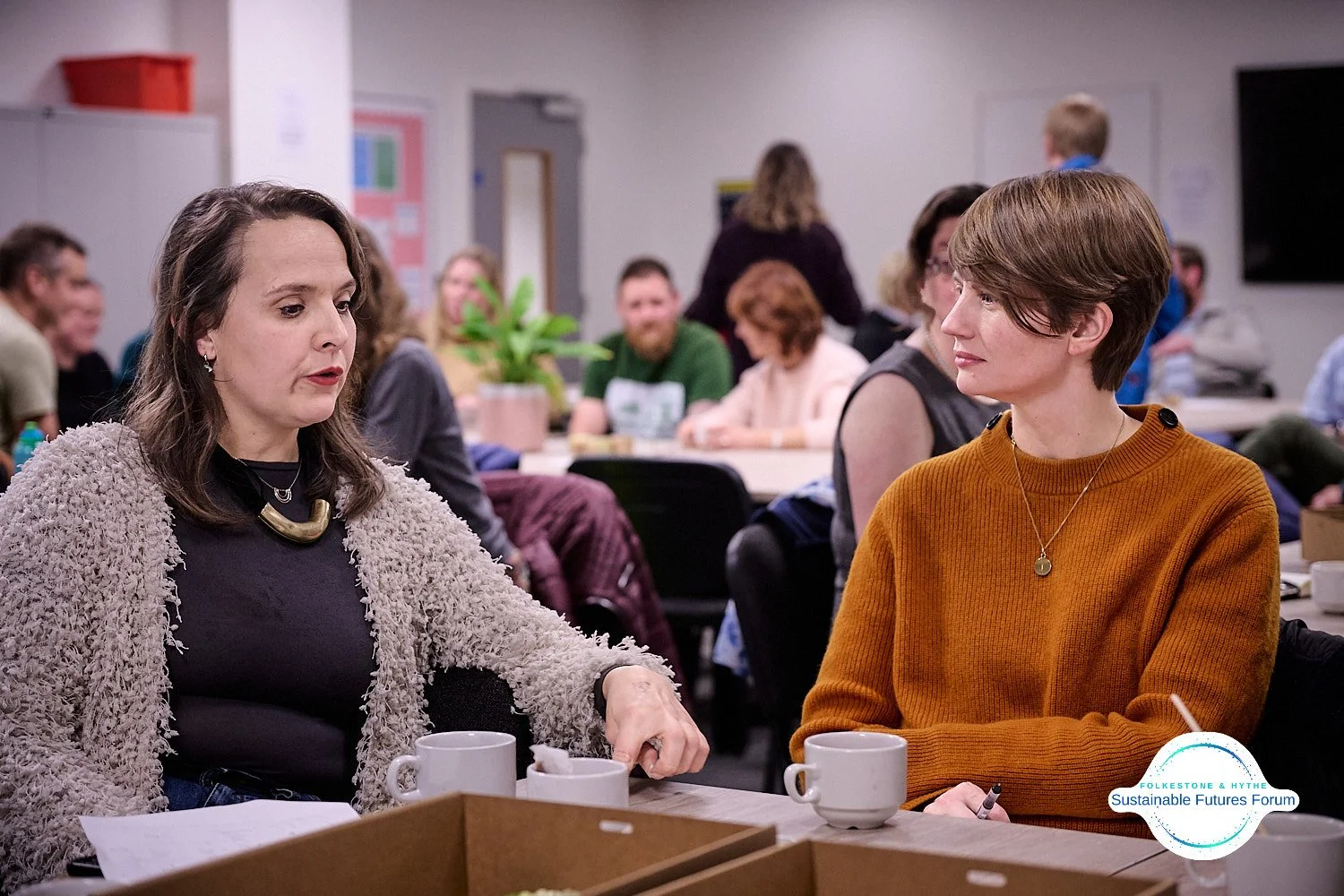 Two women engaged in conversation at a round table during a meeting or discussion, with other people in the background.