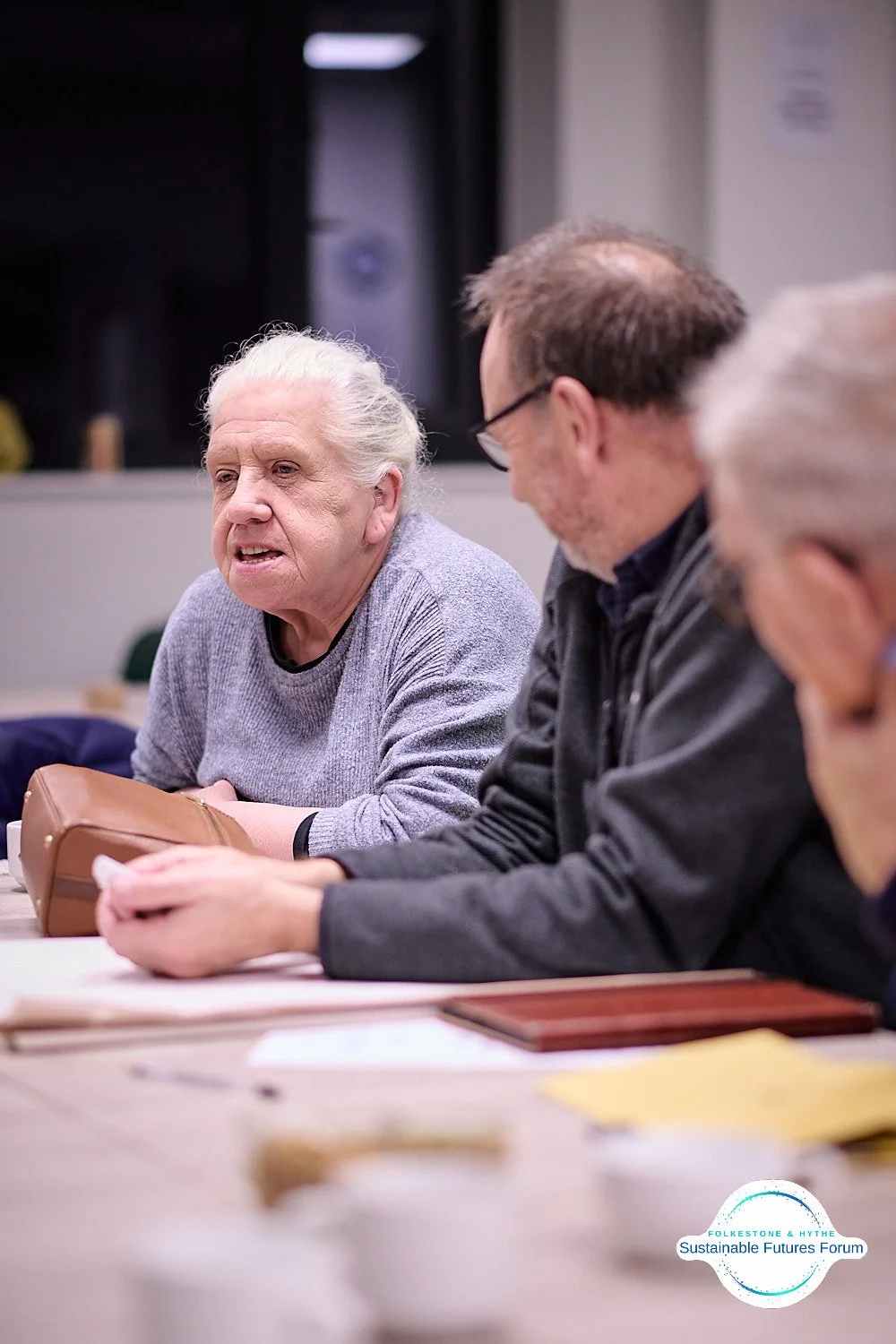 Four people sitting at a table having a discussion, with papers and folders in front of them. One older woman with white hair is speaking, while the others listen attentively. A logo in the bottom right corner reads 'Folketone & Rythe Sustainable Fut