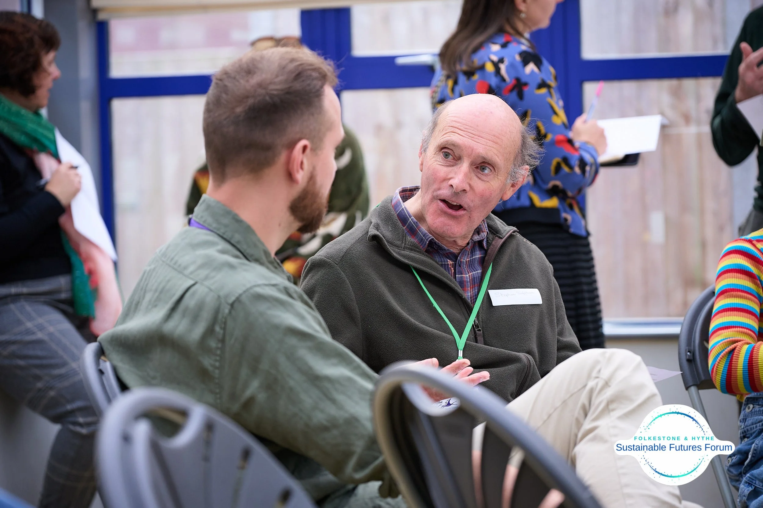 Two men engaged in conversation at the Sustainable Futures Forum, with others standing in the background. The man on the right has a bald head and is wearing a dark jacket, and the man on the left has short hair and a beard, wearing a green shirt. Se