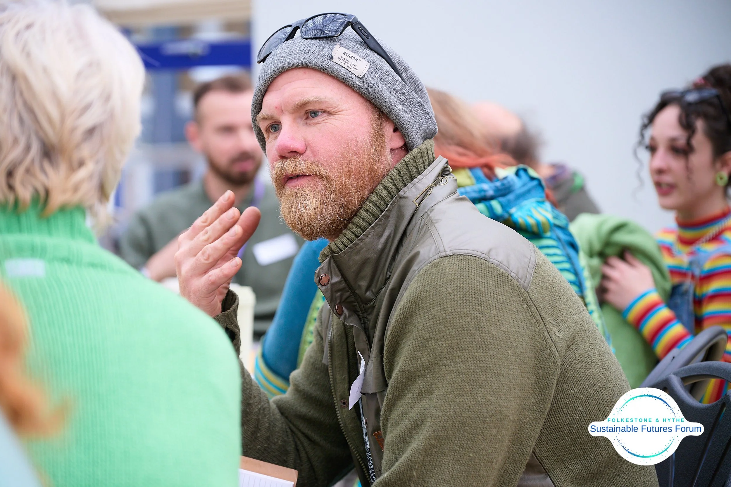 A man with a beard wearing a gray beanie and sunglasses talking to a woman in a green sweater at a forum. Several people are in the background. The logo of Folkes Tone & Hythe Sustainable Futures Forum is visible in the bottom right corner.