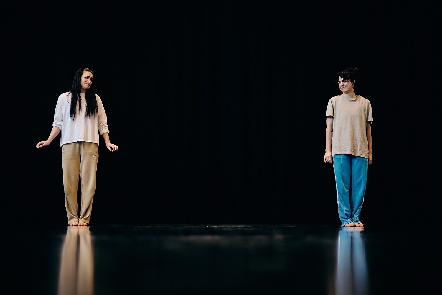 Two women standing apart on a dark stage, looking at each other, with reflections on the shiny floor.