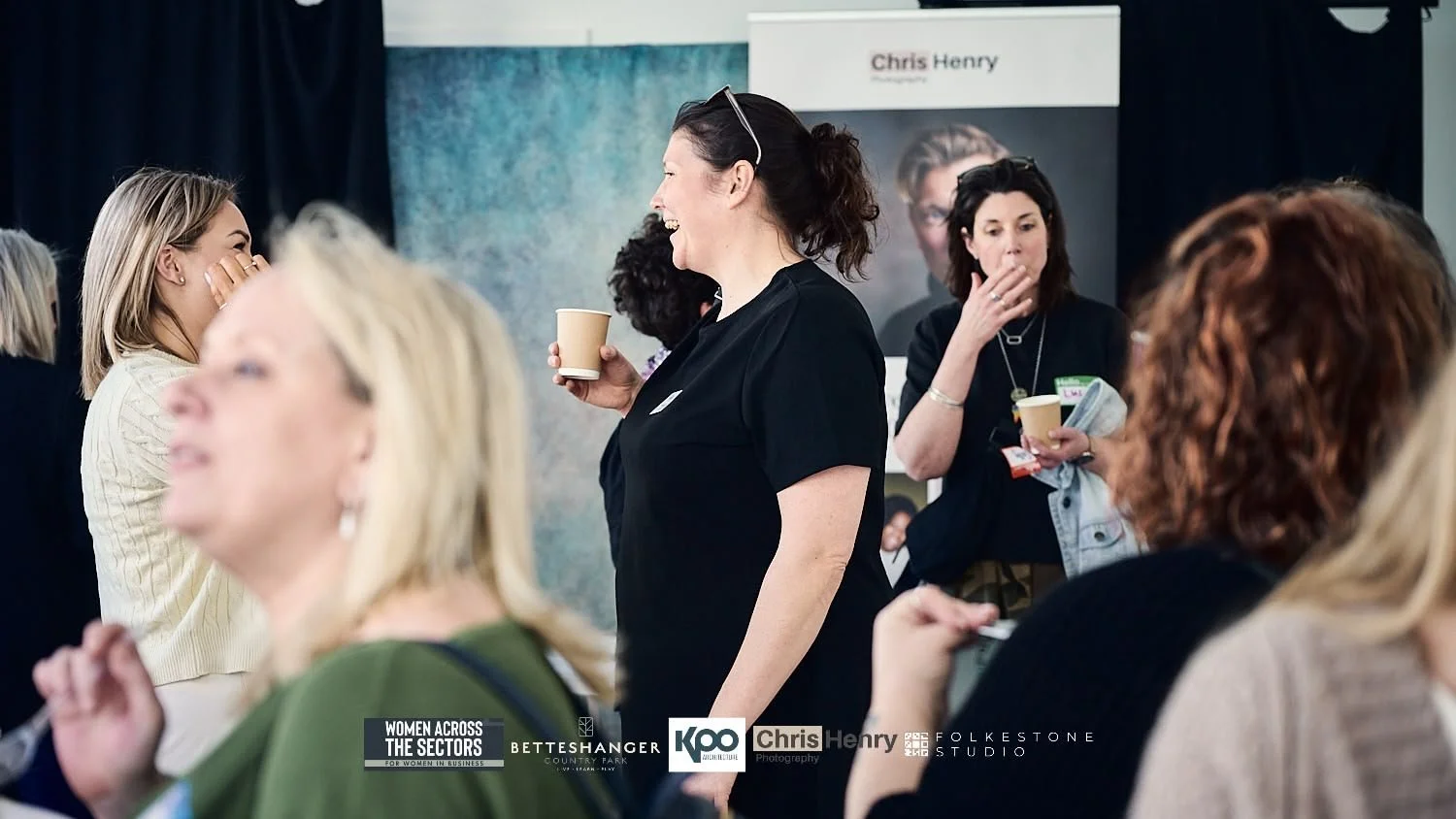 Women laughing and socializing at an event with banners and various people in attendance.