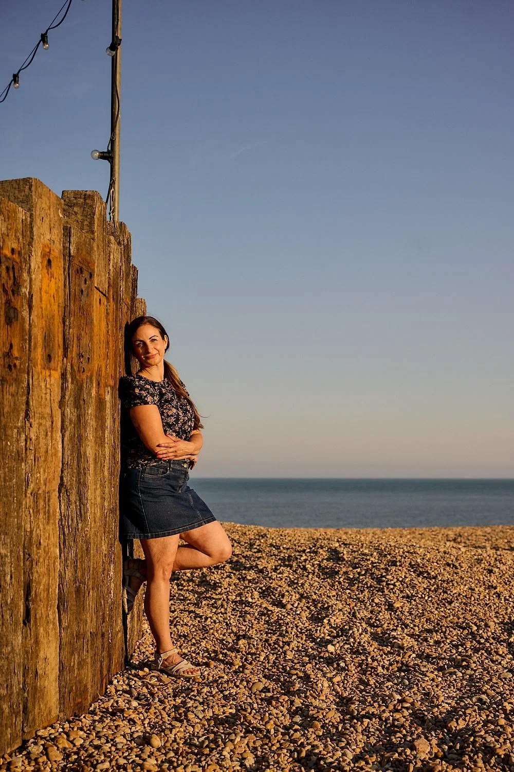 A woman leaning against a wooden post on a pebble beach, with the ocean and sky in the background during sunset.