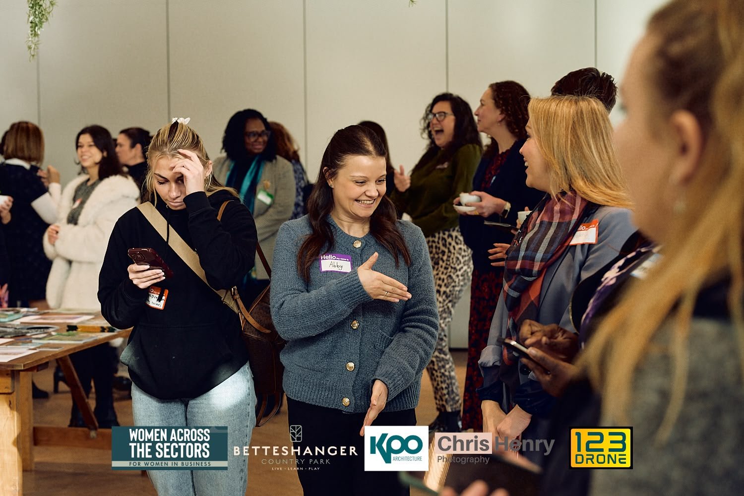Group of women at a networking event, some smiling and talking, others engaged with phones, in an indoor setting.