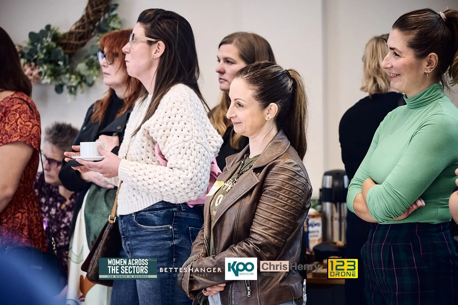 A group of women standing in a line at an indoor event, listening attentively and smiling, with some holding cups, in a professional or networking setting.