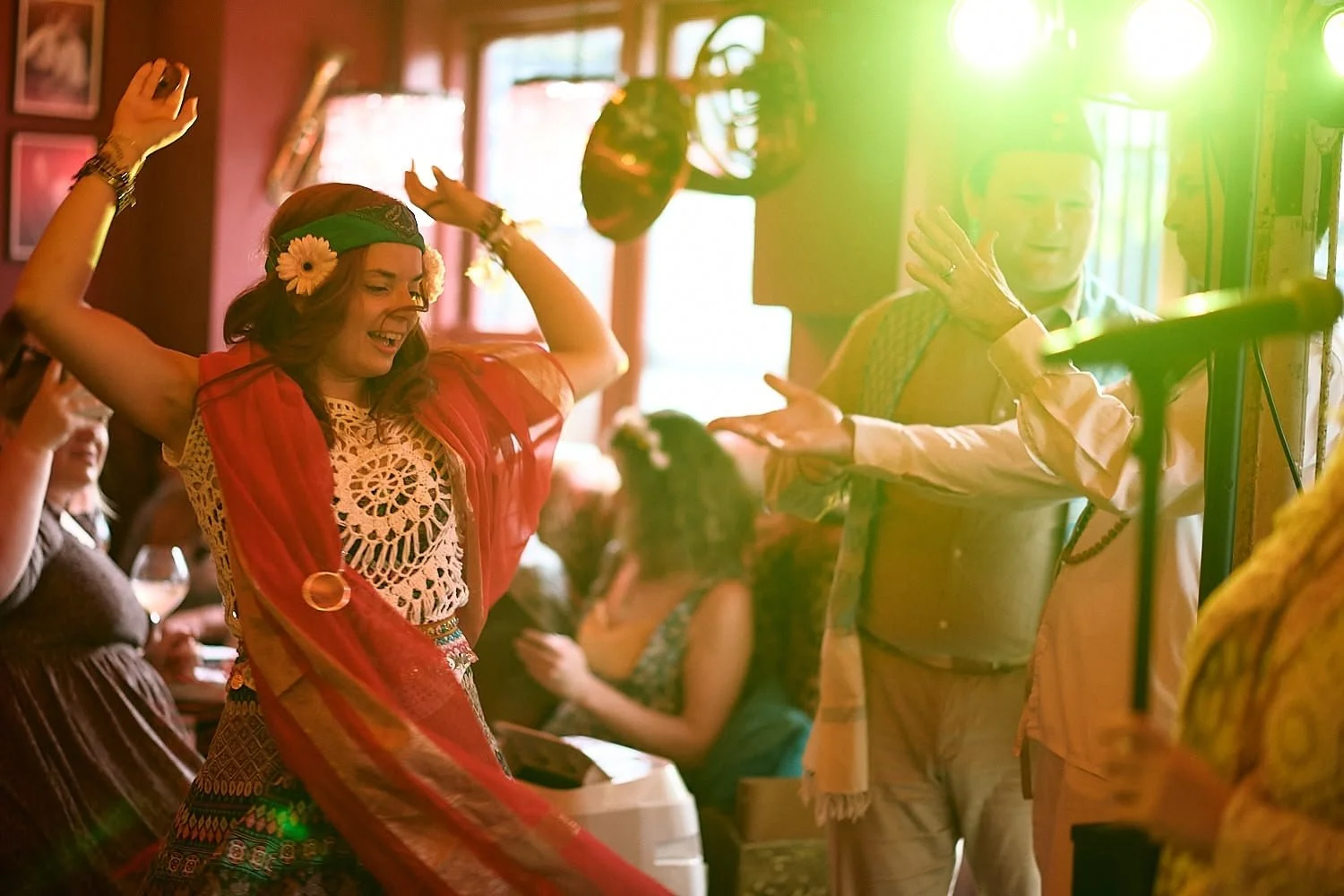 A woman dancing joyfully with her arms raised, wearing a cloth headband with flowers, in a lively indoor setting with other people in the background. A person nearby is playing a musical instrument.