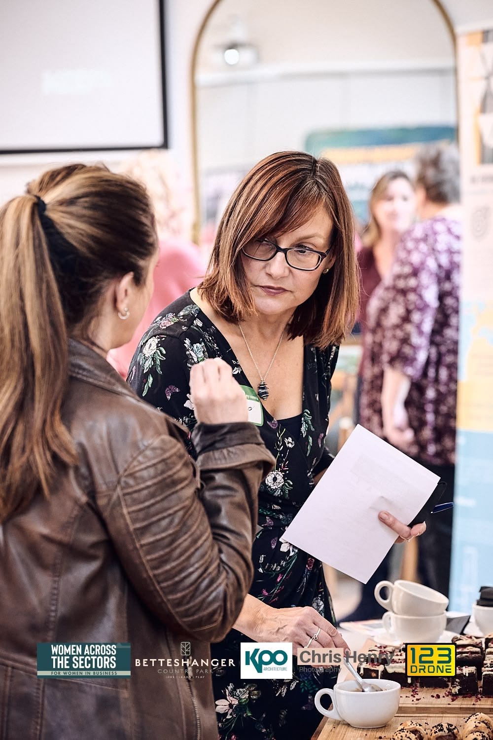 Two women are engaged in conversation at an event, with one holding a napkin and the other gesturing with her hand. There are coffee cups and a plate of cookies on the table nearby. The background shows other attendees and various informational poste