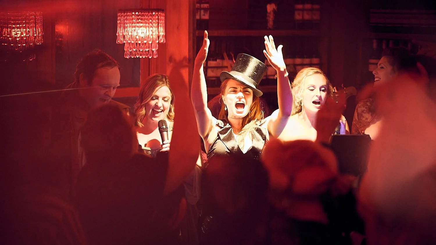 A group of people singing and dancing at a lively party or karaoke event, with one woman wearing a top hat and singing passionately with arms raised.