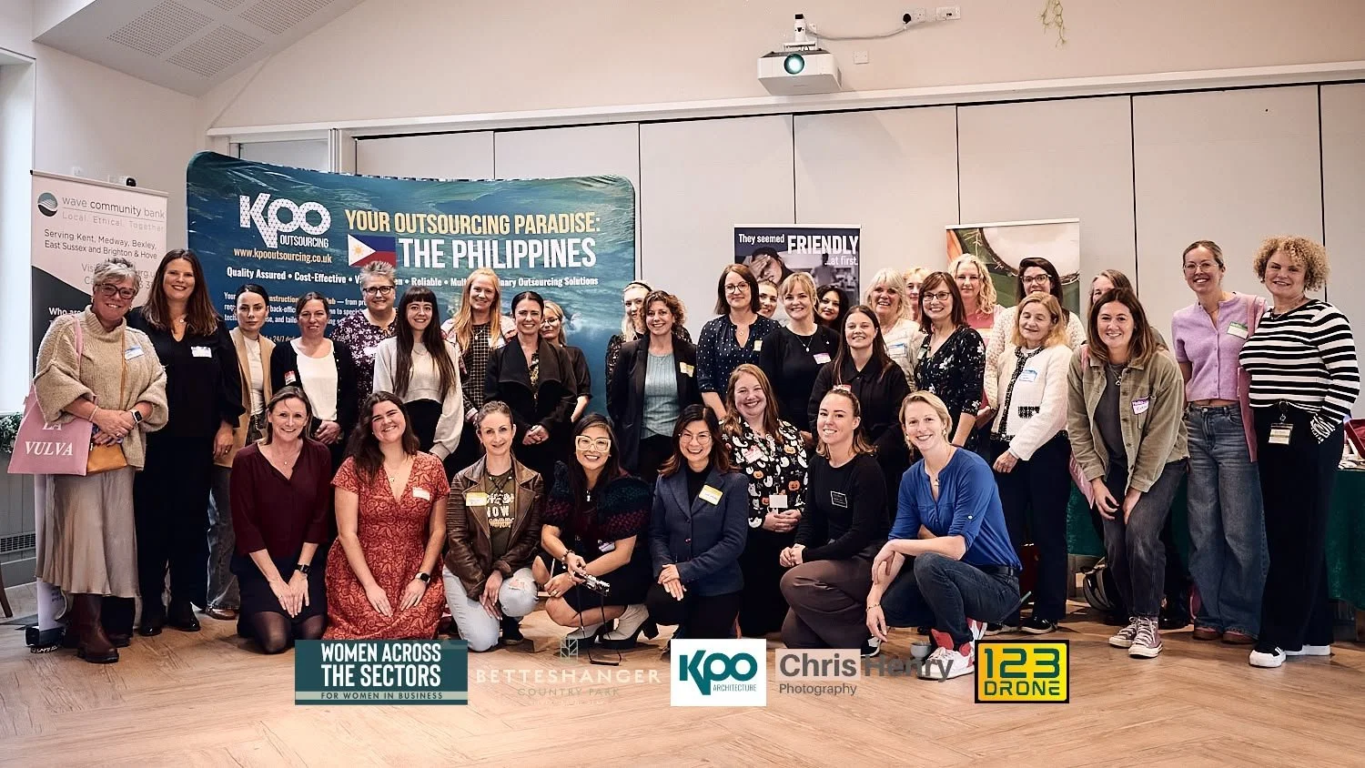 Group of women at a conference or event, standing and sitting, smiling for a group photo in front of a blue banner that reads 'Your Outsourcing Paradise: The Philippines.' Some people are holding cups, and there are various logos at the bottom of the