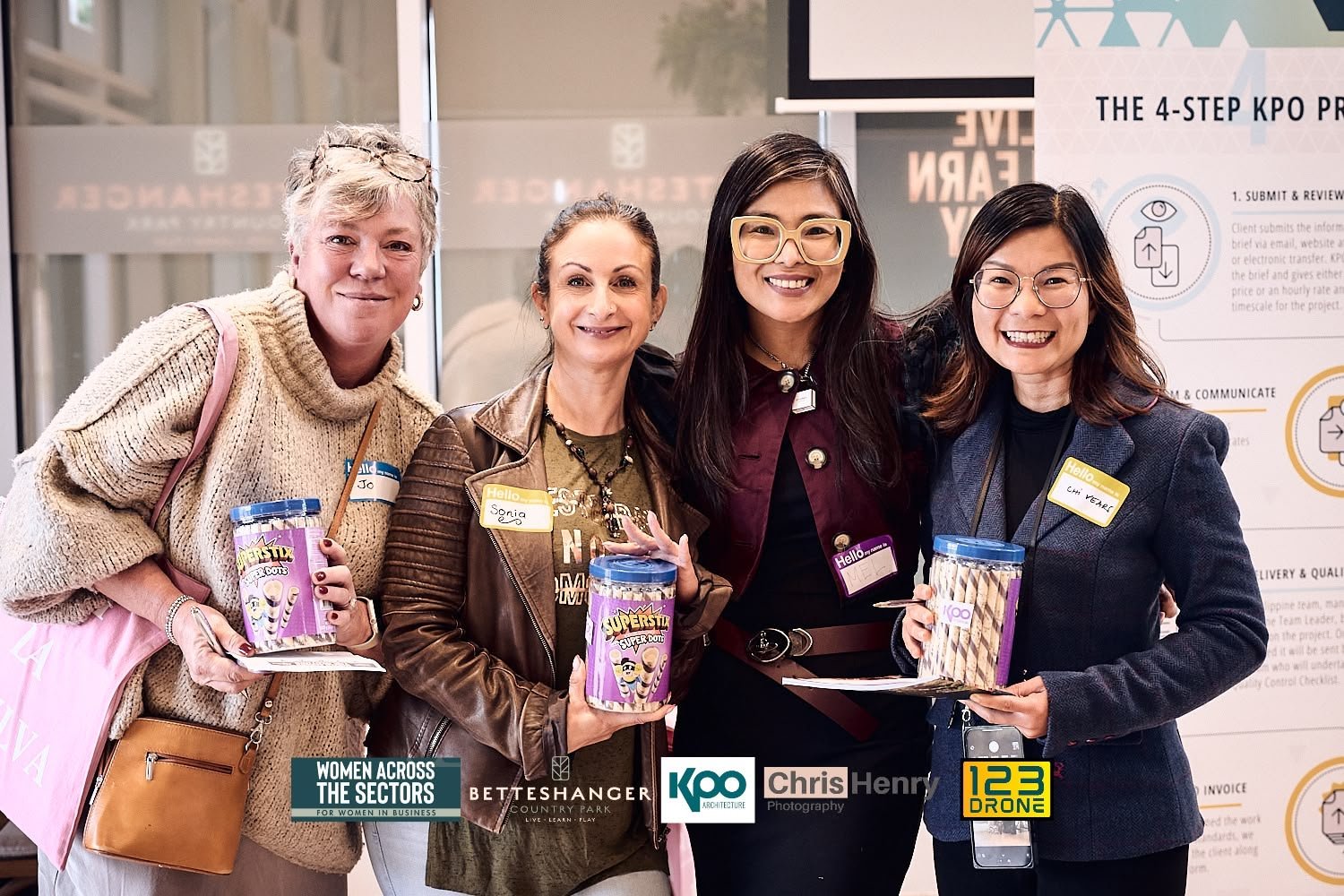 Four women standing together at a business event, smiling, holding cans of snacks, and wearing name tags.