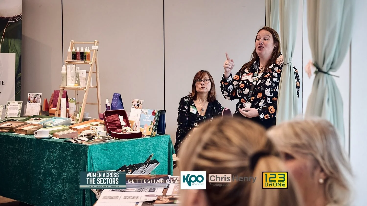 Two women speaking at a women in business event with a table of products and promotional materials in the background, and audience members in the foreground.