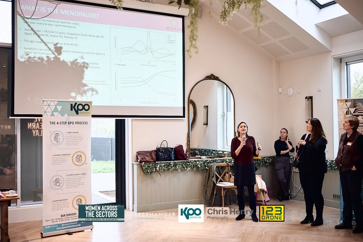 A woman is giving a presentation on menopause to a small audience in a well-lit room with large windows. A large screen displays slides with charts and information. There are four women standing and listening, with some taking notes or observing atte