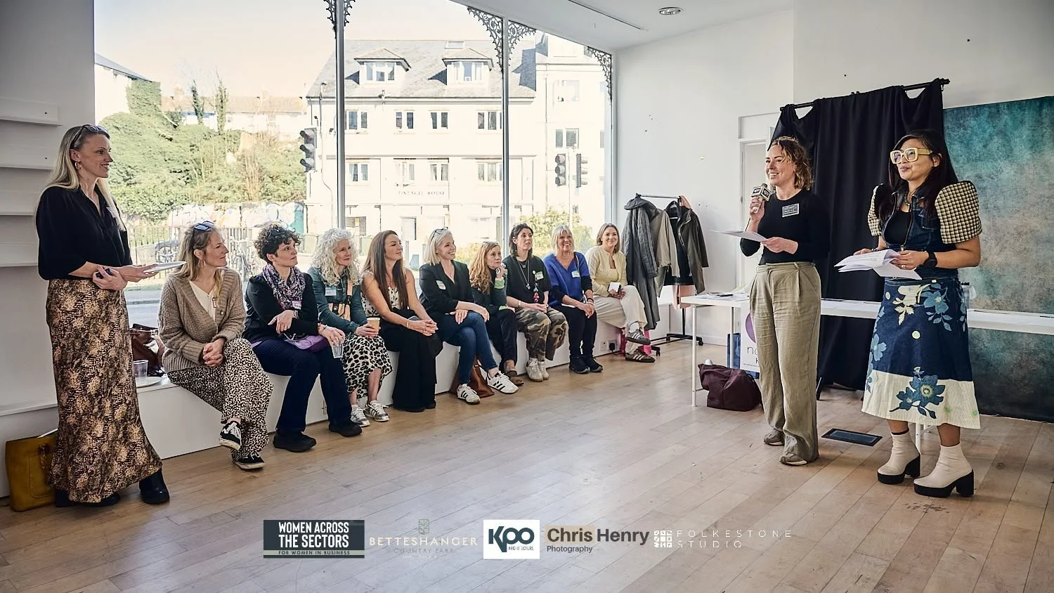 Women attending a presentation or panel discussion in a bright room with large window, some women sitting on a window seat and two women standing at the front speaking, holding papers and a microphone.