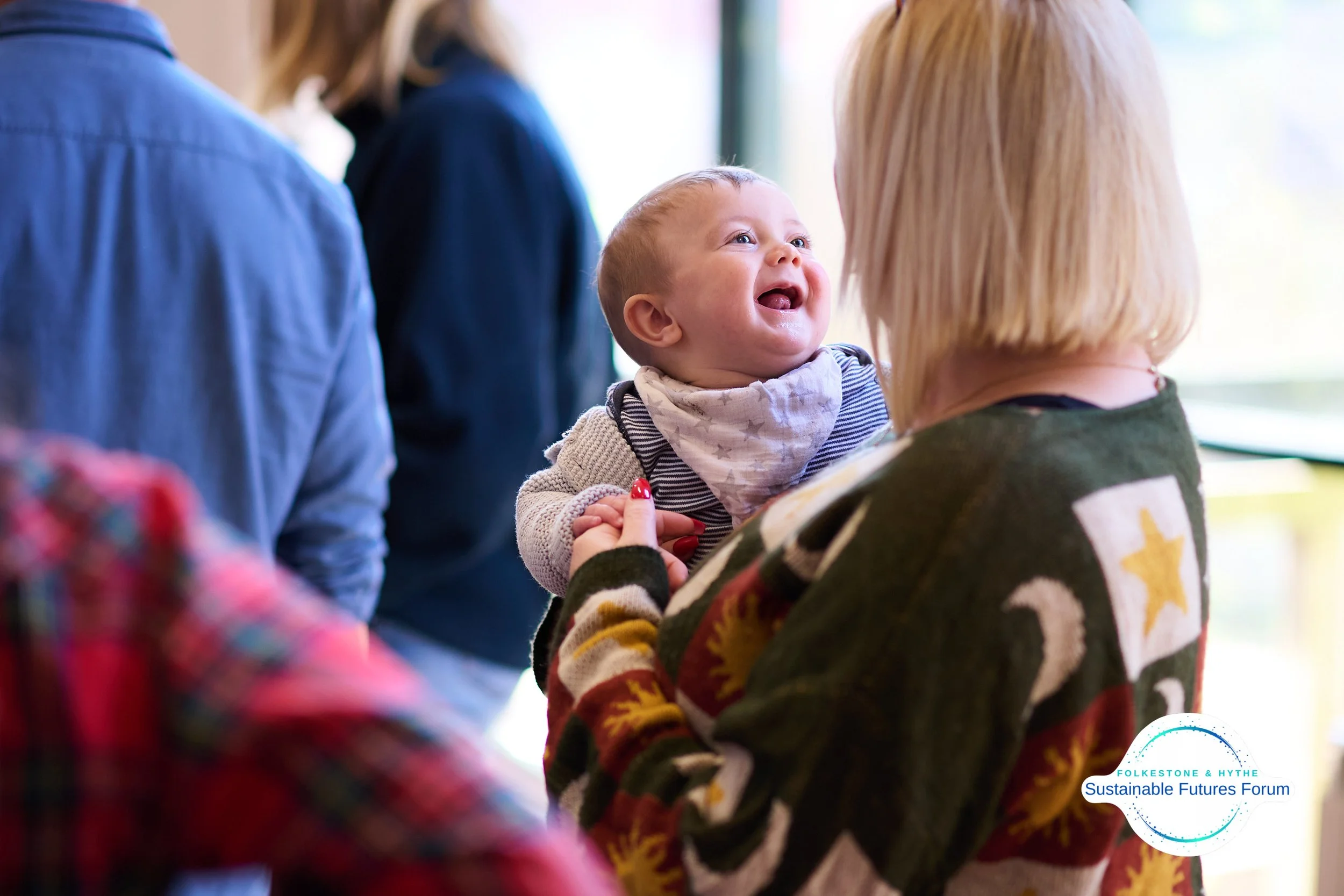 A woman holding a smiling baby with an expressive face, looking at her, in a brightly lit indoor setting with other people in the background.