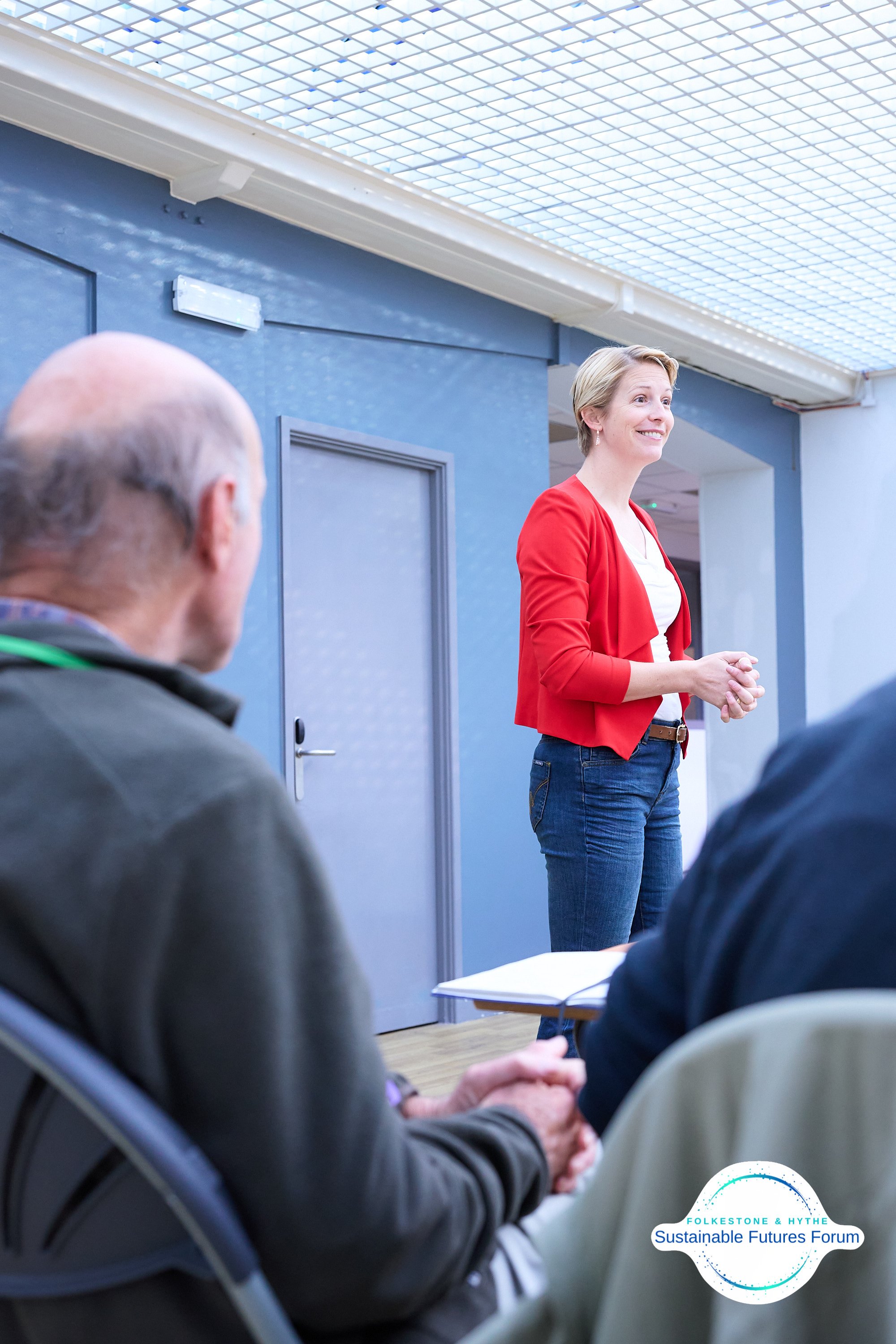 A woman in a red blazer giving a presentation to an audience in a conference room, with a blue wall and ceiling lights, featuring the Folkestone & Hythe Sustainable Futures Forum logo.