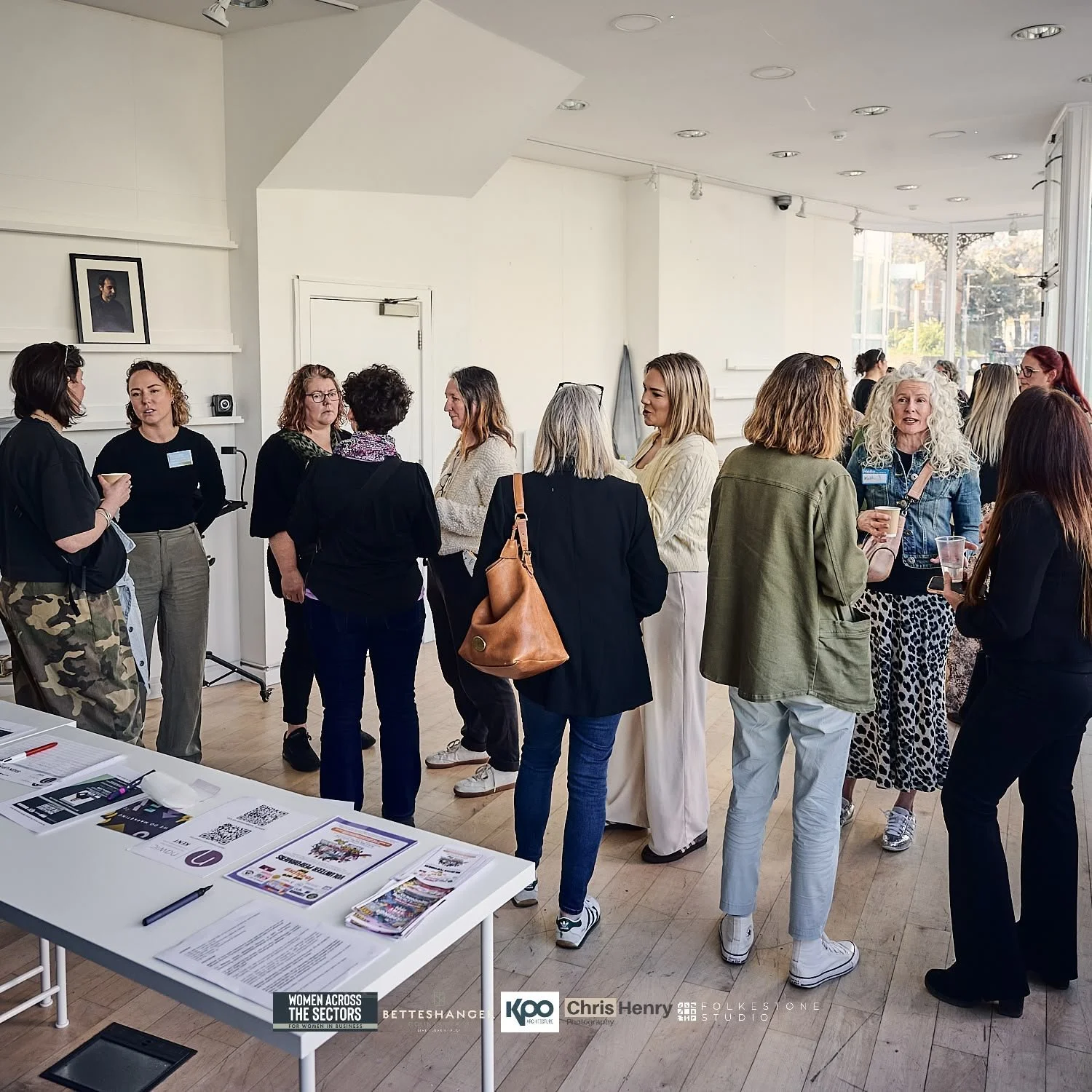 A group of women standing and talking inside a bright room with large windows, some holding cups, with a table of flyers and brochures in the foreground.