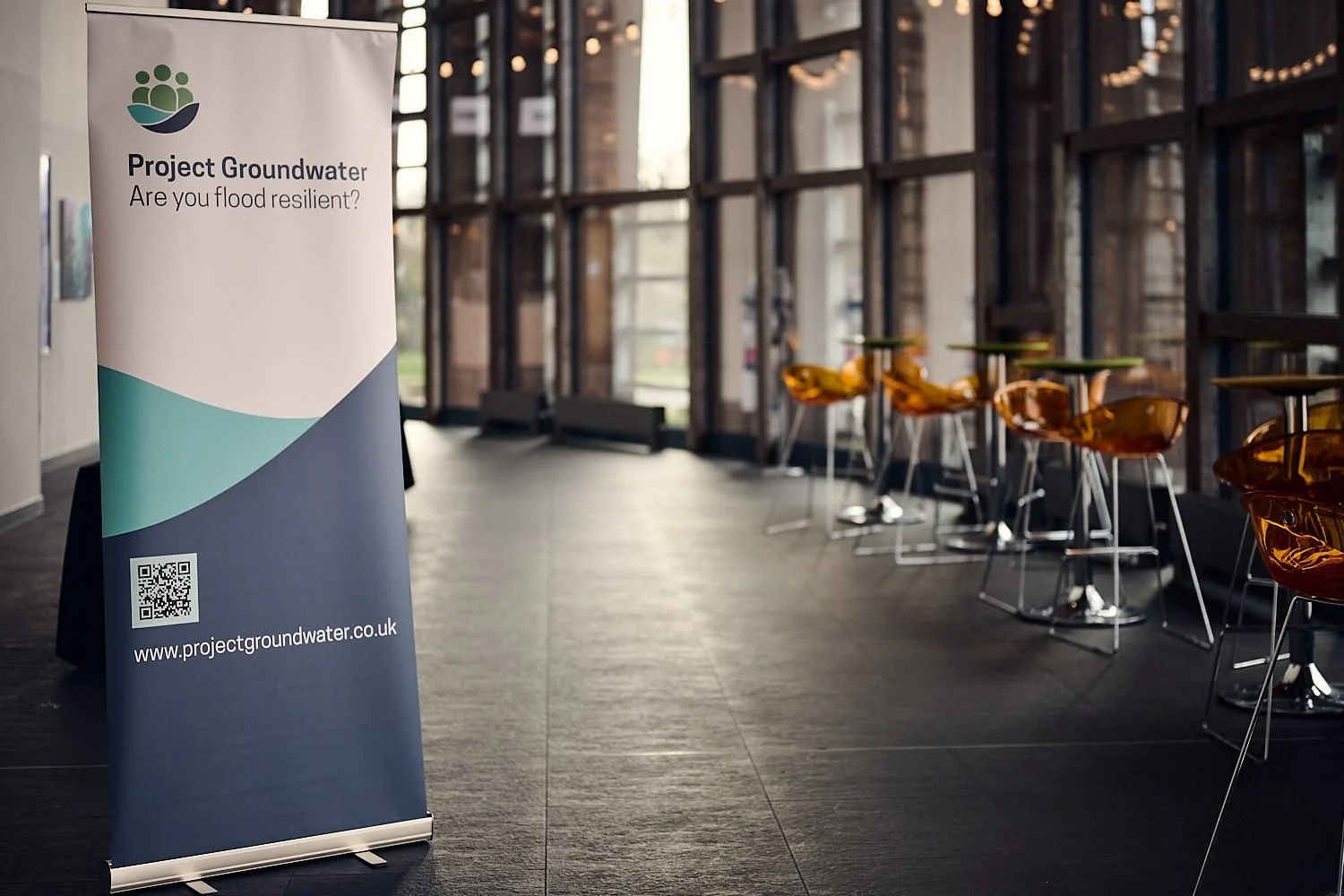 A vertical banner promoting Project Groundwater, asking if the viewer is flood resilient, with a QR code and website URL, inside a room with tall windows, black flooring, and orange chairs.